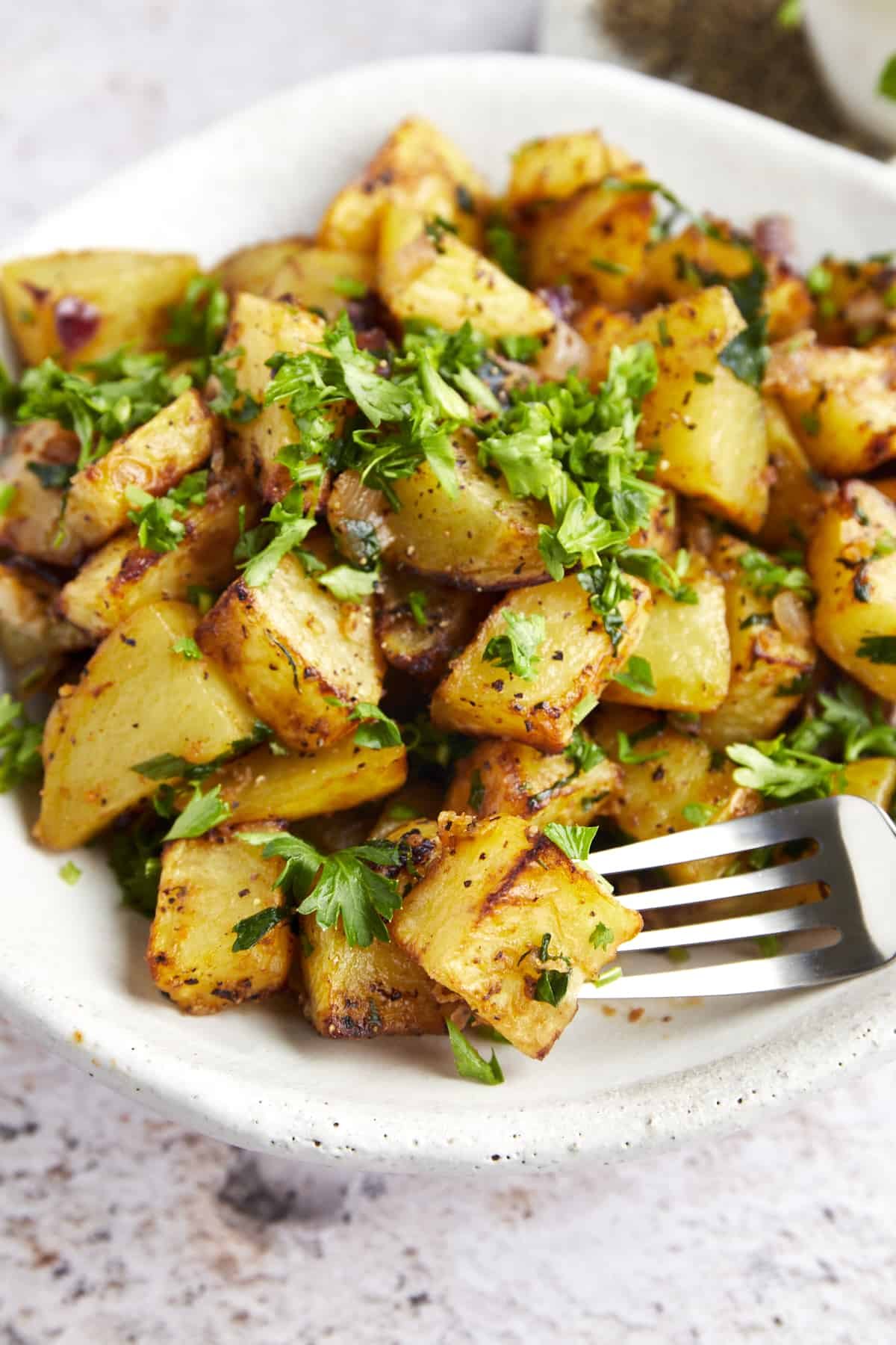 A fork with a bite of air fryer golden potato next to a plate full of potatoes.