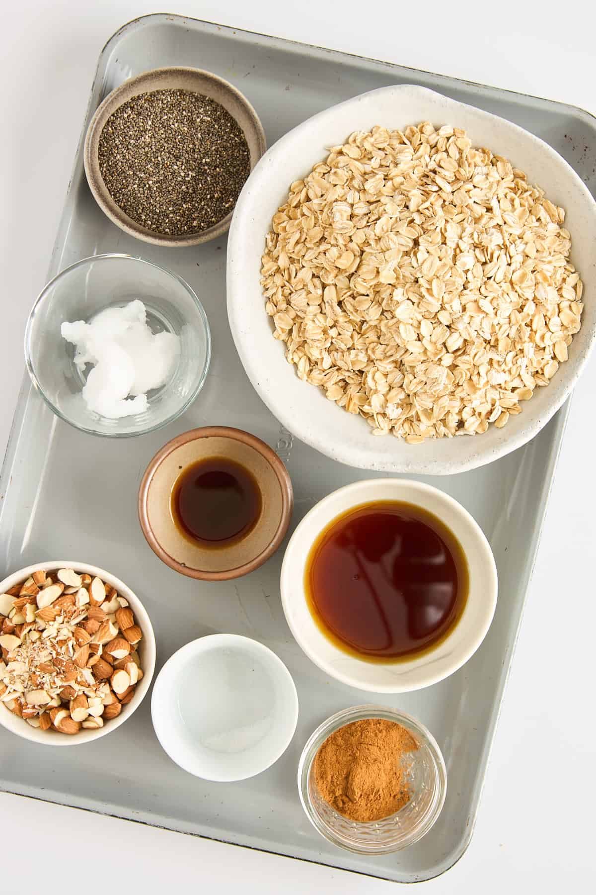 A tray with bowls containing rolled oats, chia seeds, coconut oil, chopped almonds, vanilla extract, maple syrup, salt, and ground cinnamon arranged neatly for baking.