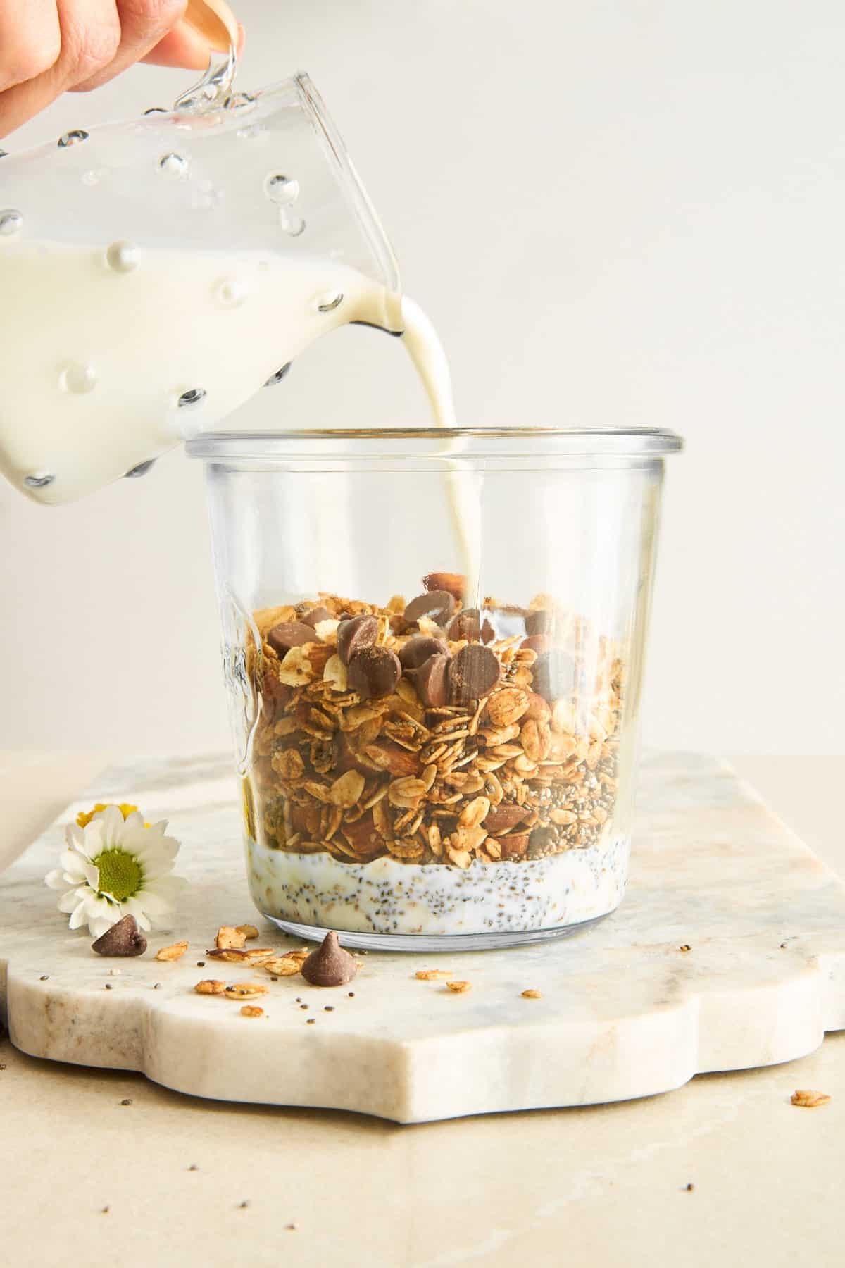 A hand pours milk from a glass pitcher into a clear jar filled with granola, chocolate chips, and chia seeds, sitting on a marble board with a small white flower nearby.