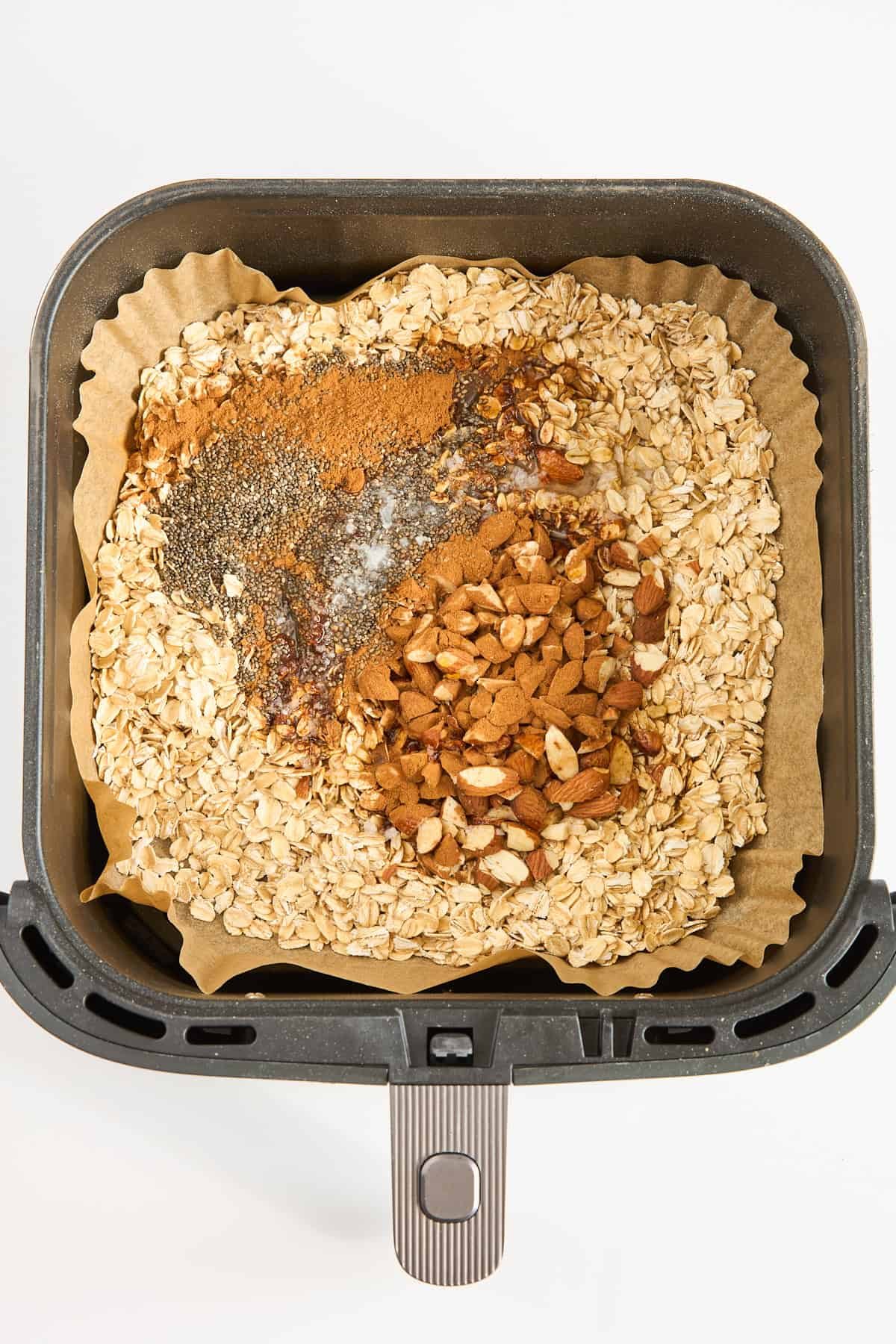 An air fryer basket lined with parchment paper contains dry oats, chopped nuts, chia seeds, cinnamon, and a drizzle of honey, ready to be mixed.