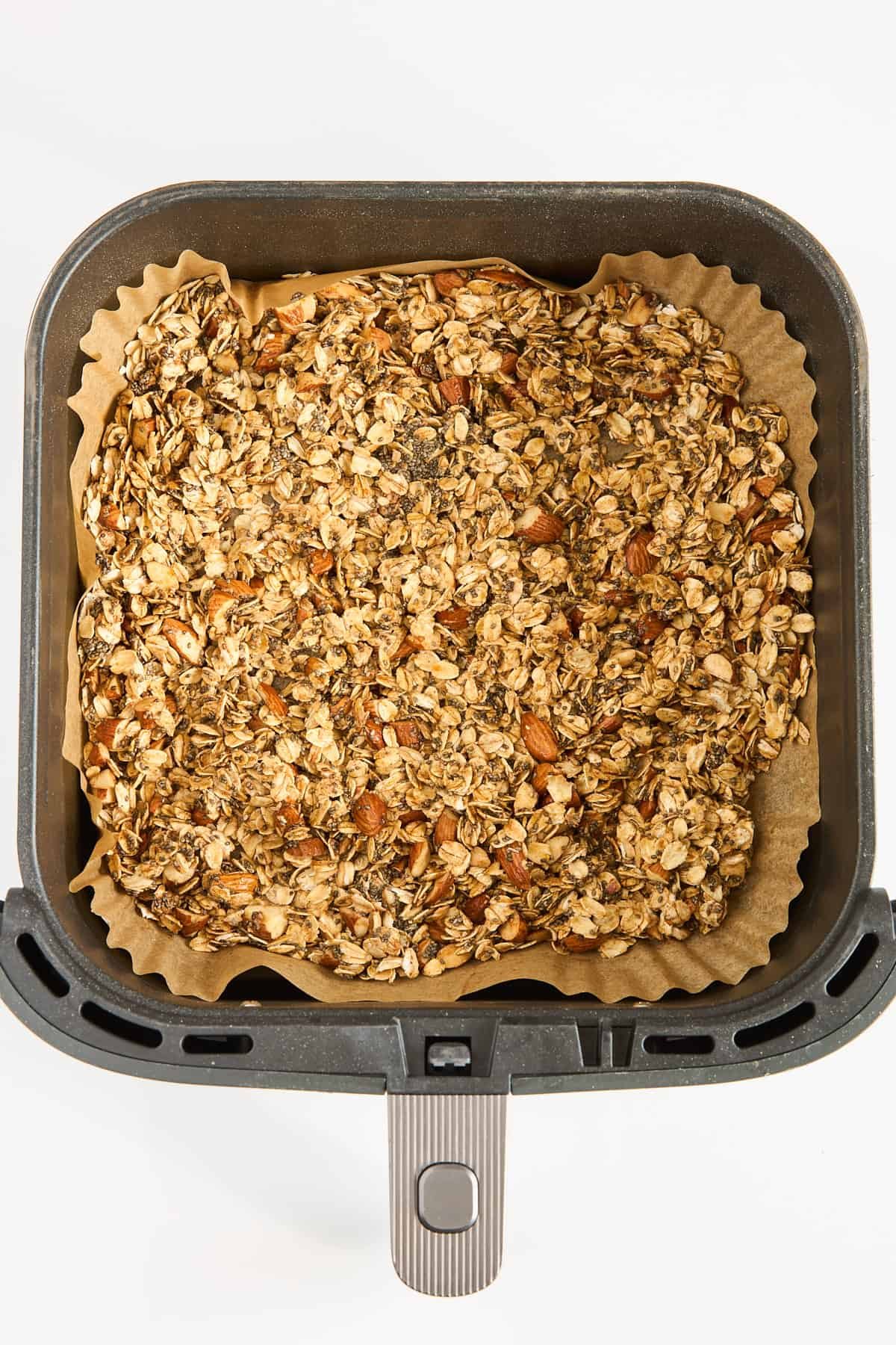 A square air fryer basket lined with parchment paper contains an even layer of granola with visible oats and almonds, ready for baking.
