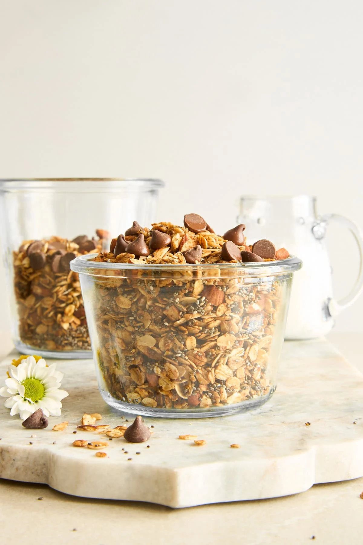 Two glass jars filled with granola and chocolate chips sit on a light stone surface. A small white flower and chocolate chips are scattered nearby. A glass pitcher of milk is in the background.