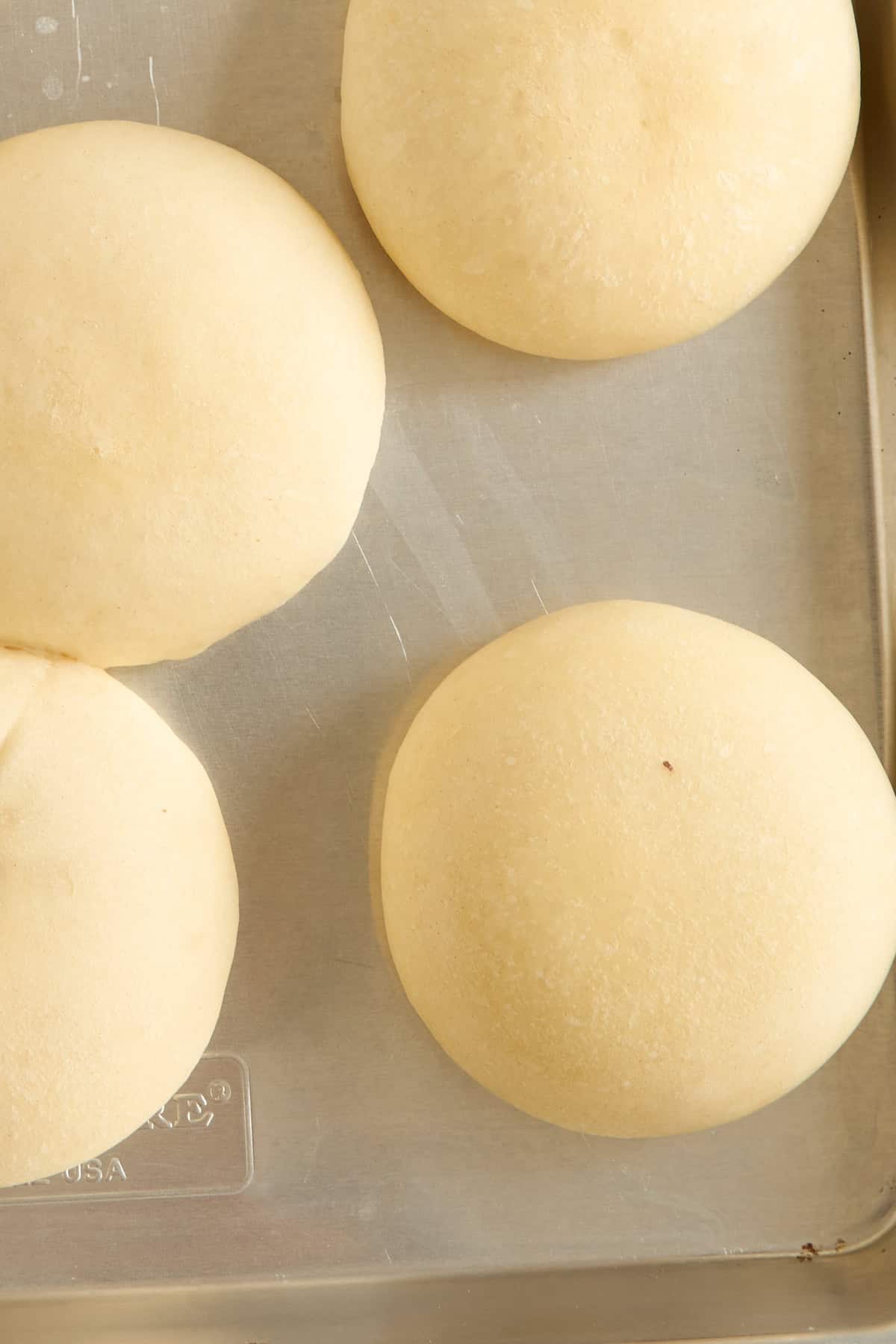 Four round, unbaked dough balls are arranged on a metal baking tray, ready to be baked. The surface of the dough is smooth and lightly dusted with flour.