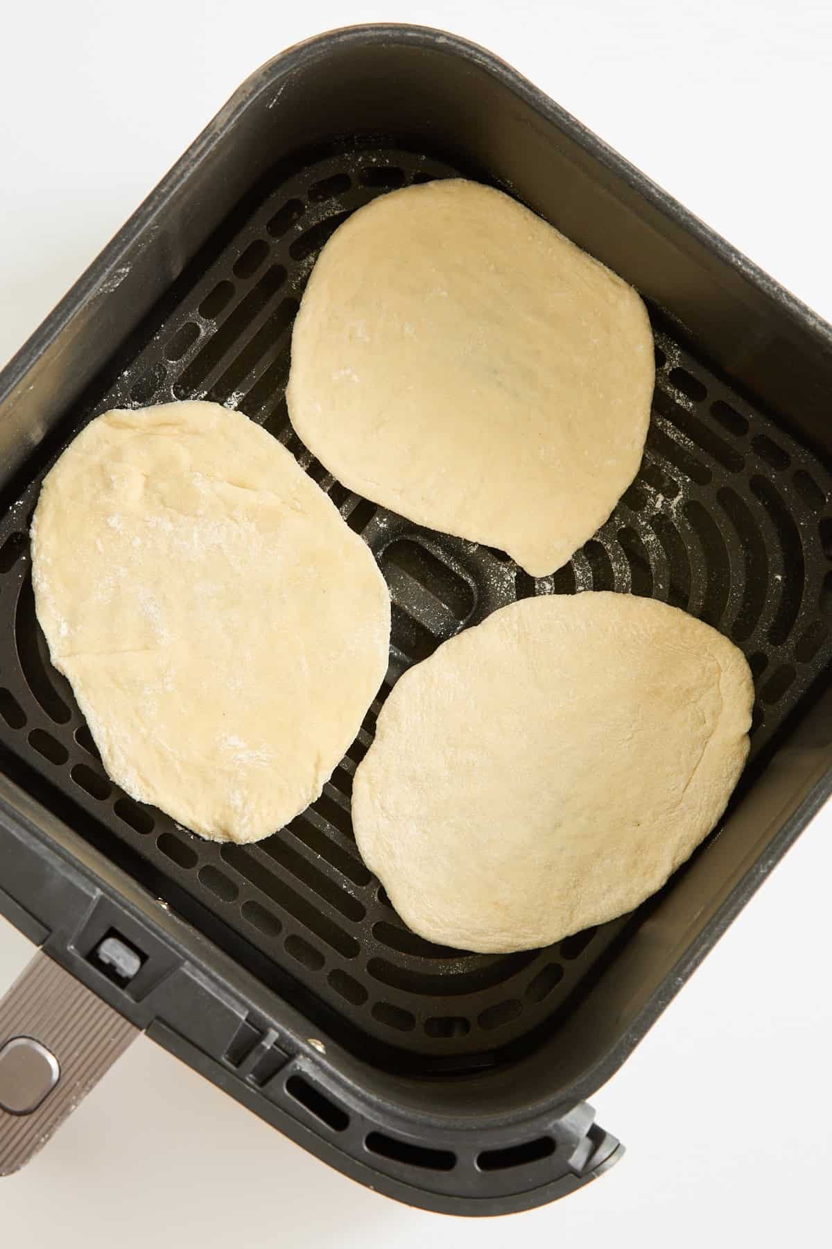 Three pieces of raw flatbread dough are placed inside the basket of an air fryer, ready to be cooked. The basket is black with a nonstick, perforated surface.