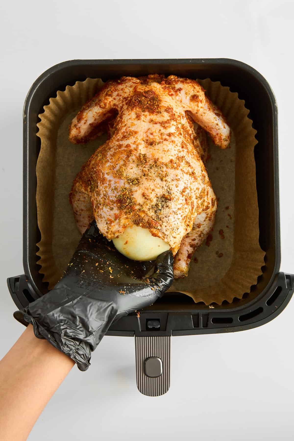 A gloved hand stuffing a raw whole chicken in an air fryer.