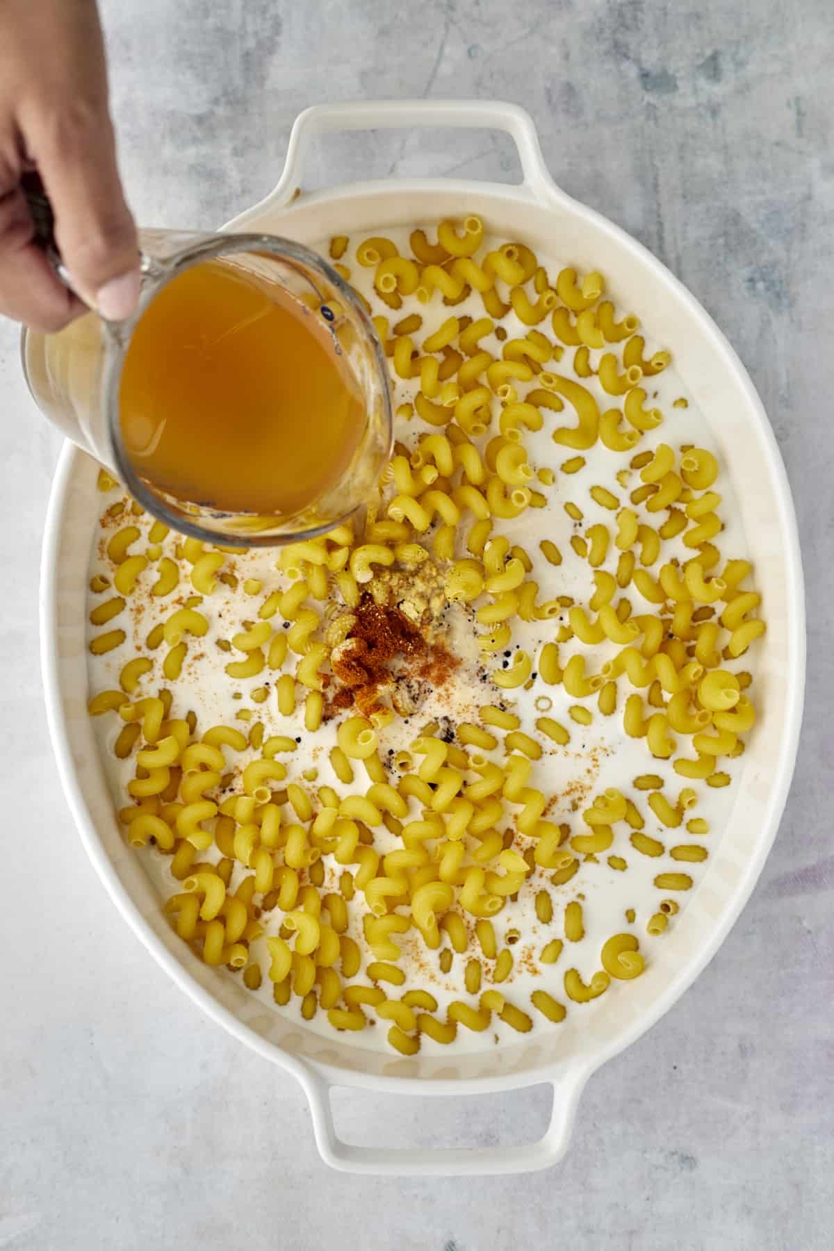 broth and spices being poured over milk and cavatappi noodles in an oval baking dish.