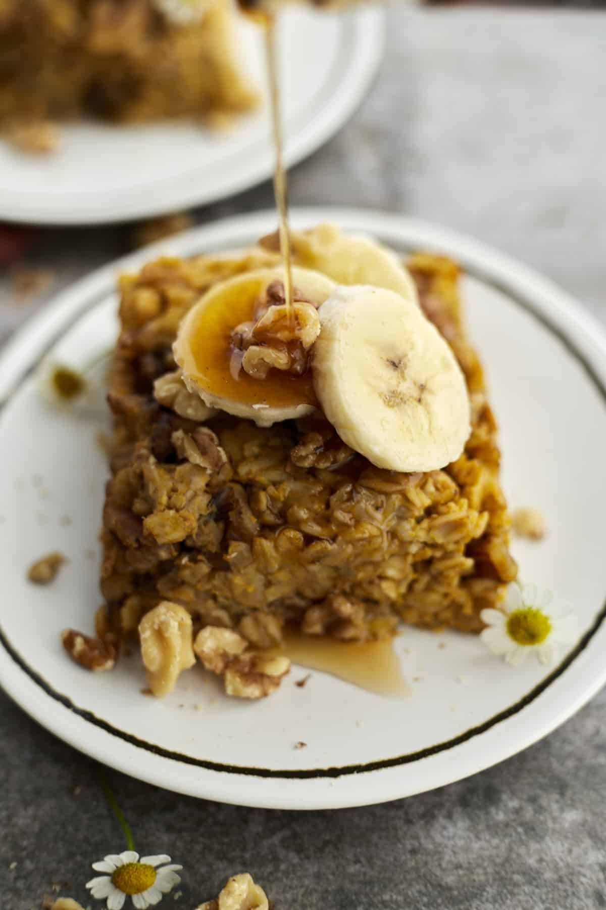 a slice of baked pumpkin oatmeal topped with walnuts and banana coins with a drizzle of syrup being poured on top