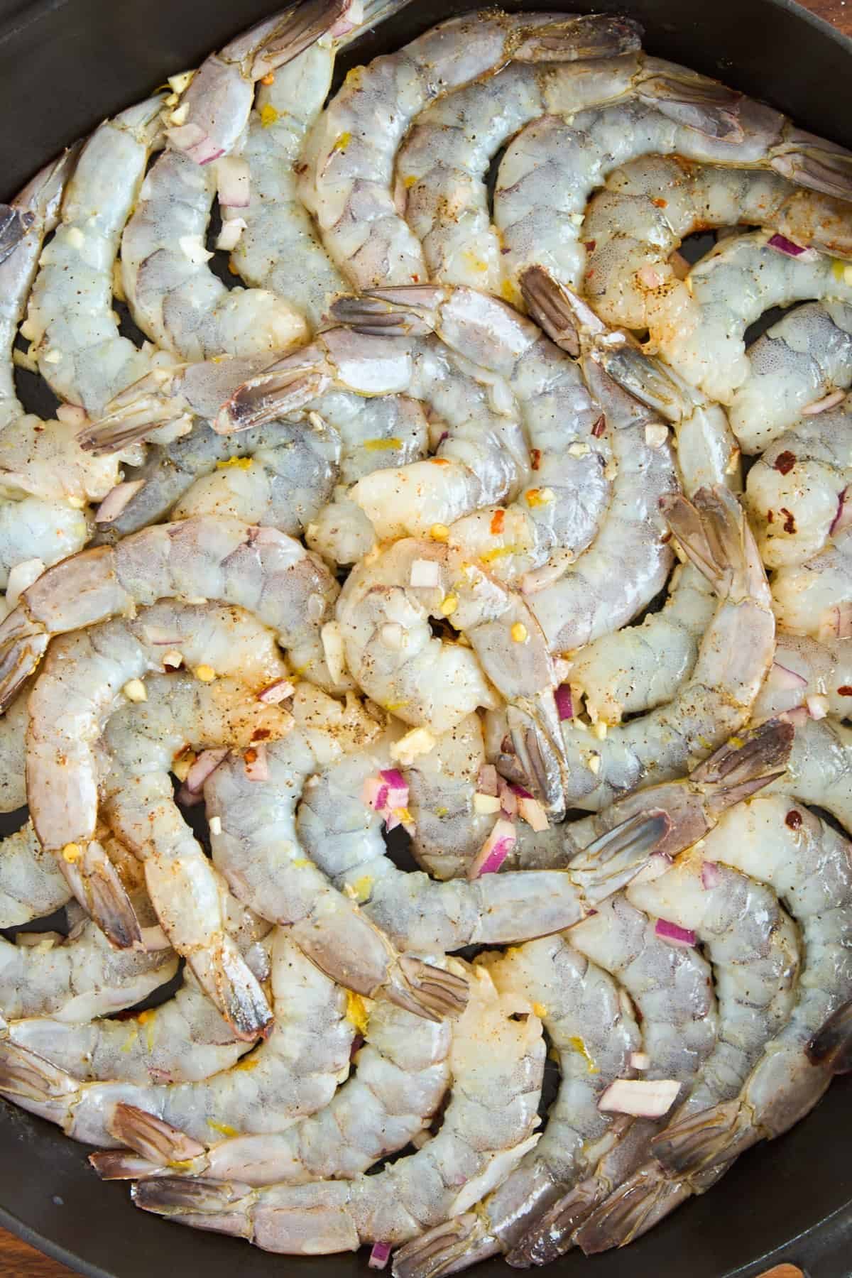 A close-up of raw shrimp arranged in a circular pattern in a pan, sprinkled with chopped garlic, red onion, and spices.