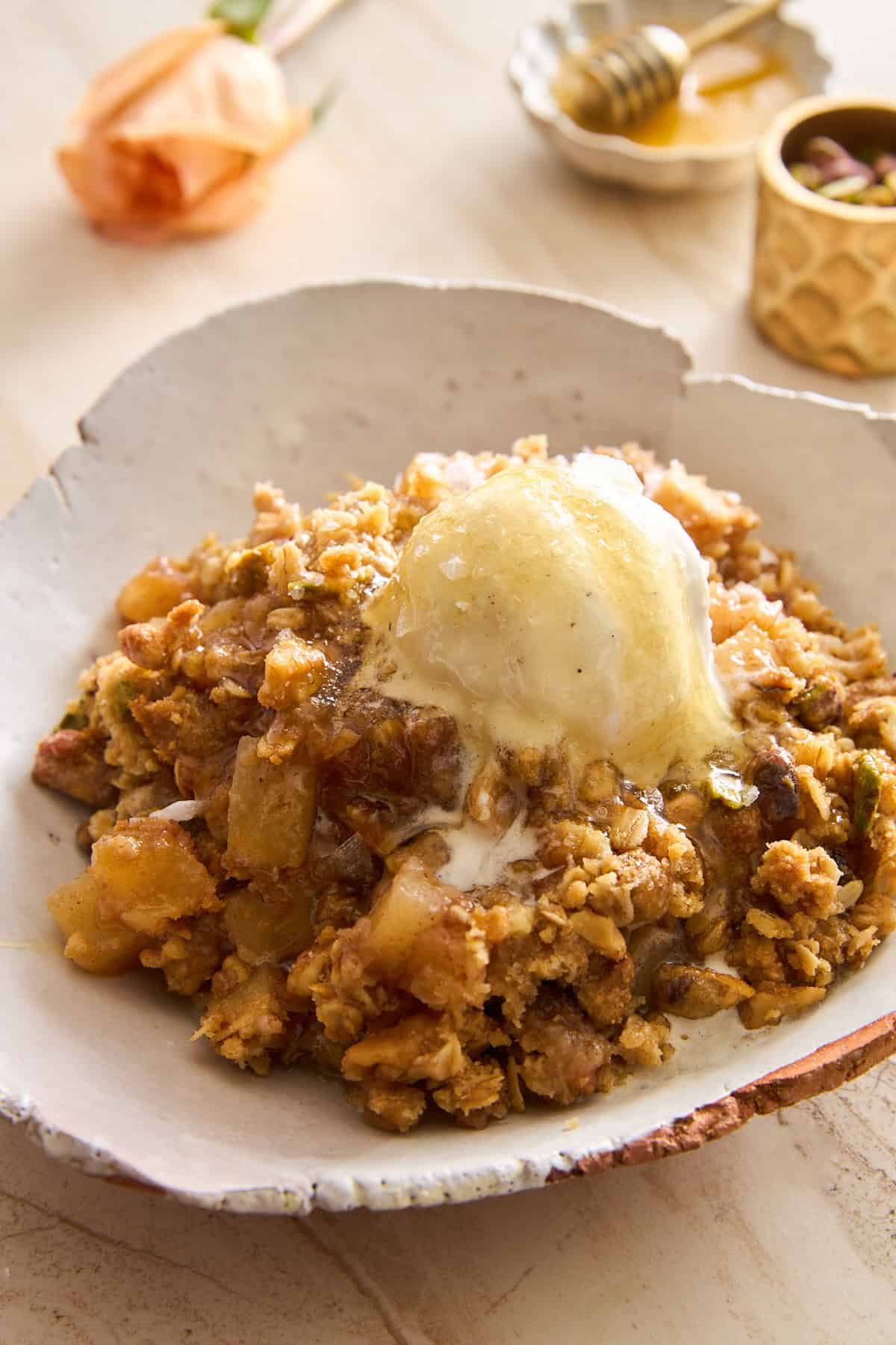 A serving of baklava apple crisp topped with a scoop of melting vanilla ice cream on a rustic ceramic plate, with a honey dipper, small dish, and flower blurred in the background.