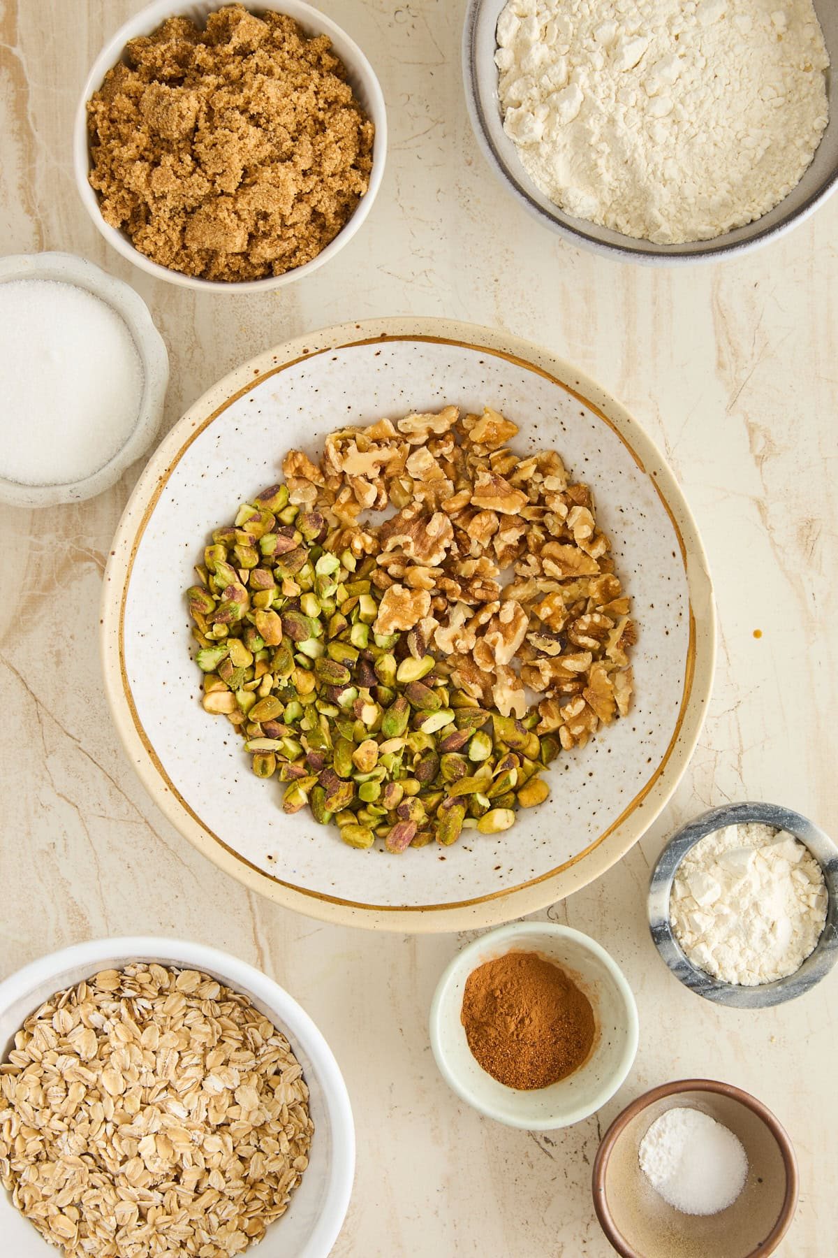 A variety of baking ingredients in bowls on a light surface, including walnuts, pistachios, brown sugar, flour, oats, cinnamon, cornstarch, salt, and granulated sugar.
