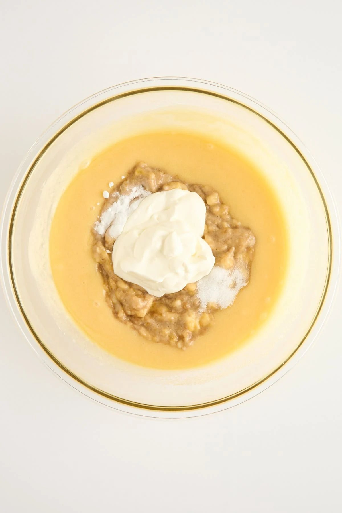 A glass bowl filled with a yellow batter, mashed bananas, white yogurt, and a sprinkle of baking soda, viewed from above on a white surface.