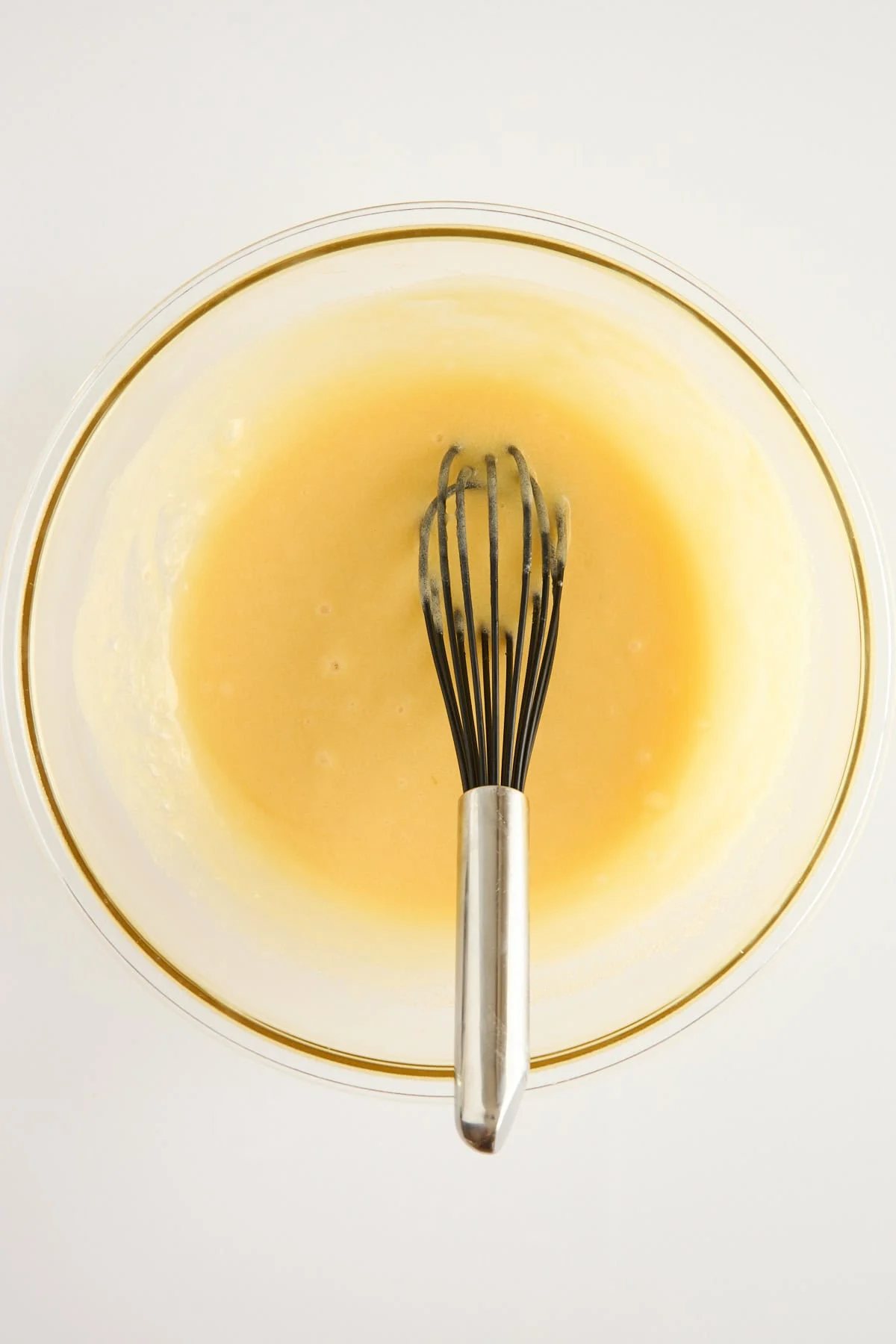A metal whisk in a glass bowl containing pale yellow batter or mixture, set against a white background.