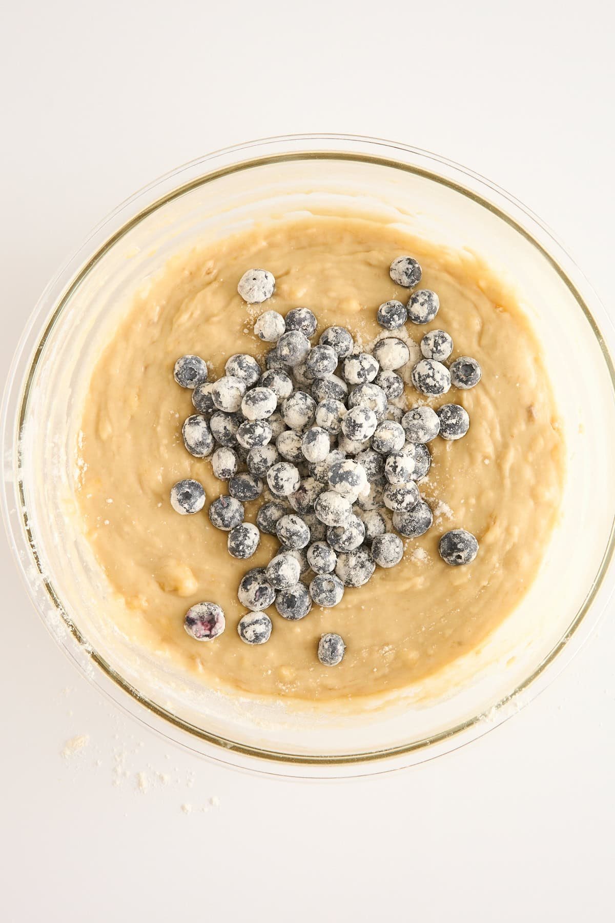 A glass bowl filled with muffin batter sits on a white surface. Fresh blueberries dusted with flour are piled on top, ready to be mixed into the batter. Some scattered flour is visible on the surface nearby.