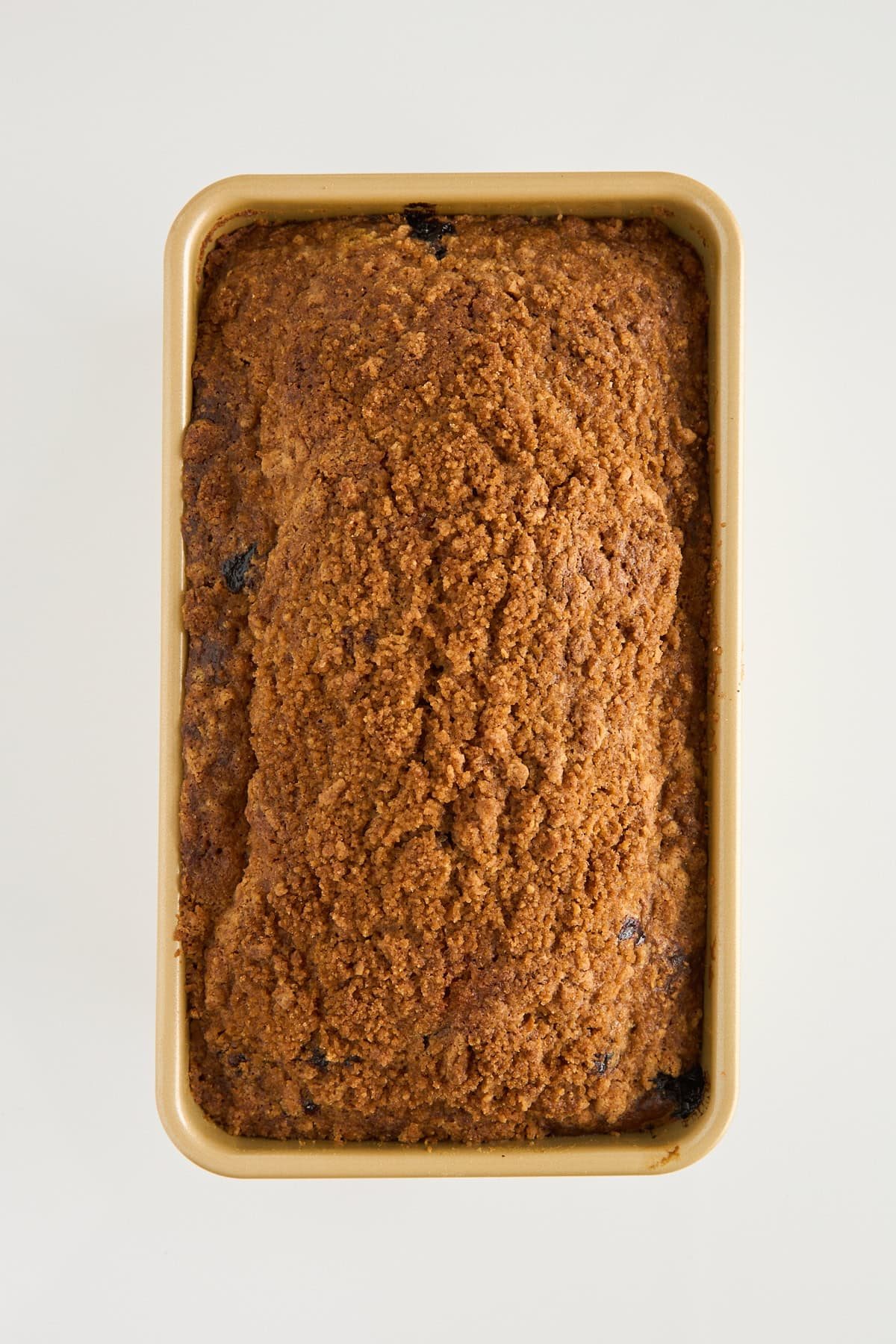 A rectangular loaf of baked bread with a crumbly brown crust is shown in a tan loaf pan, viewed from above on a white background.