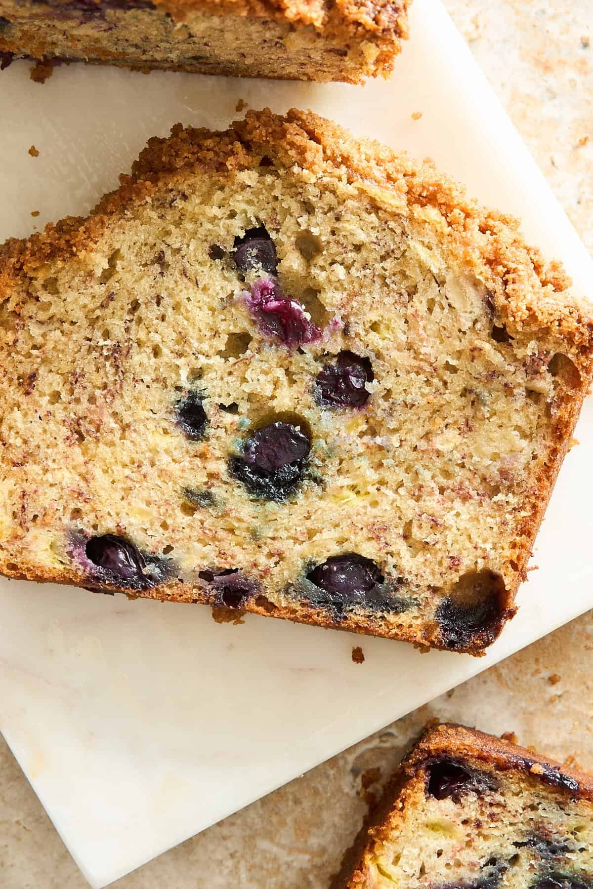 A close-up of a slice of blueberry zucchini bread on a white surface, showing a golden-brown crust and moist interior with visible blueberries and shredded zucchini.