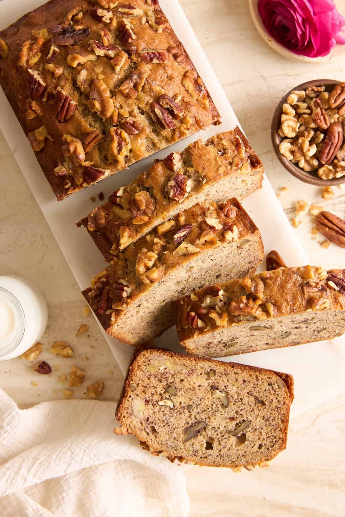 A loaf of banana nut bread, topped with chopped walnuts and pecans, sits on a white cutting board. Several slices are cut, and a bowl of mixed nuts is nearby. A glass of milk and a pink flower are also visible.