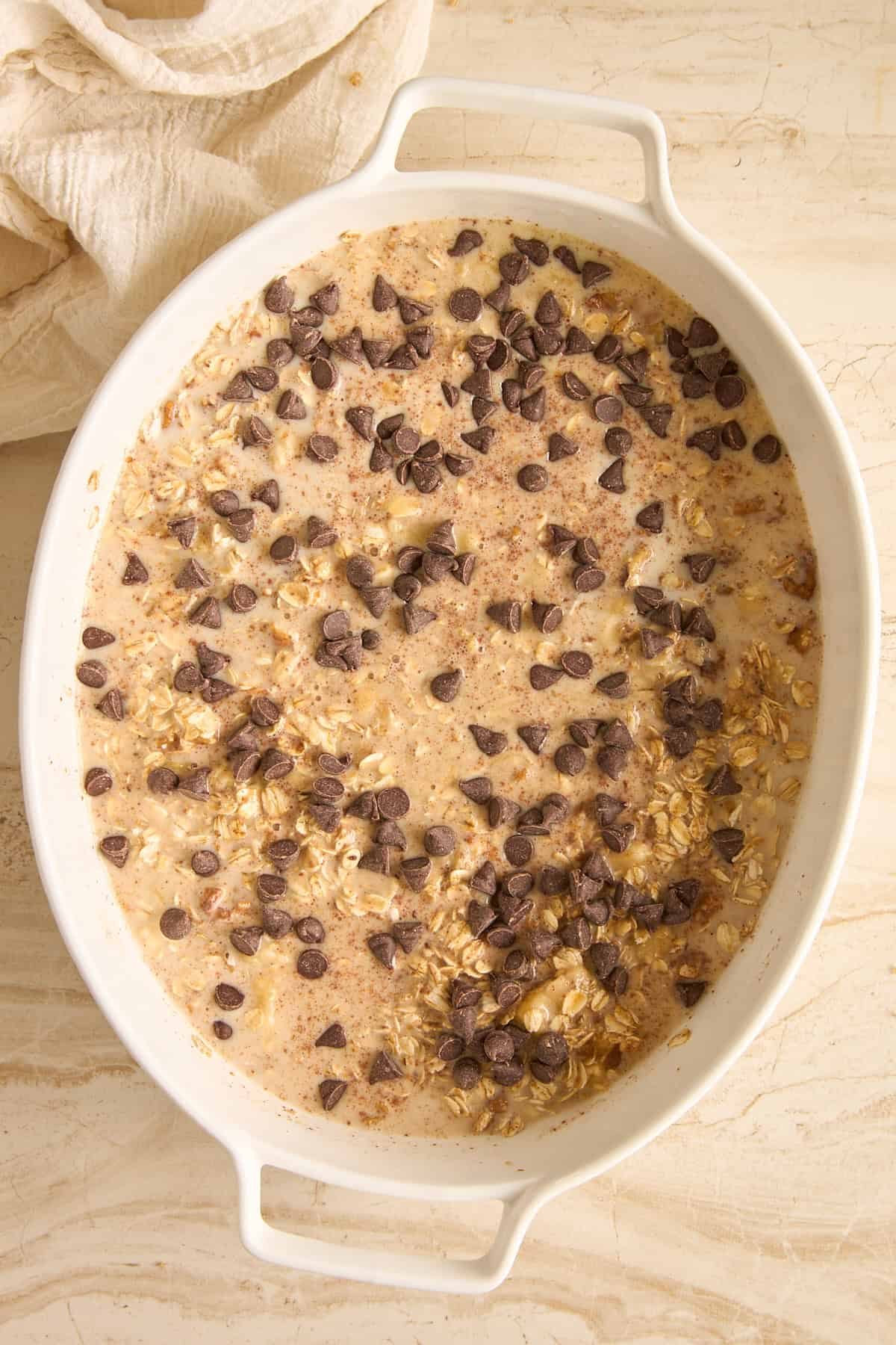 An oval white baking dish filled with uncooked oatmeal mixture, sprinkled with chocolate chips, sits on a light marble surface next to a beige cloth.