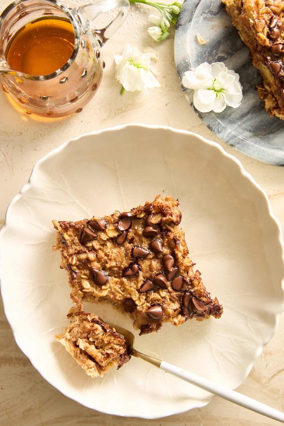 A white plate with a square slice of baked oatmeal topped with chocolate chips, a fork holding a small bite, a glass pitcher of honey, white flowers, and part of another baked oatmeal slice on a gray plate nearby.