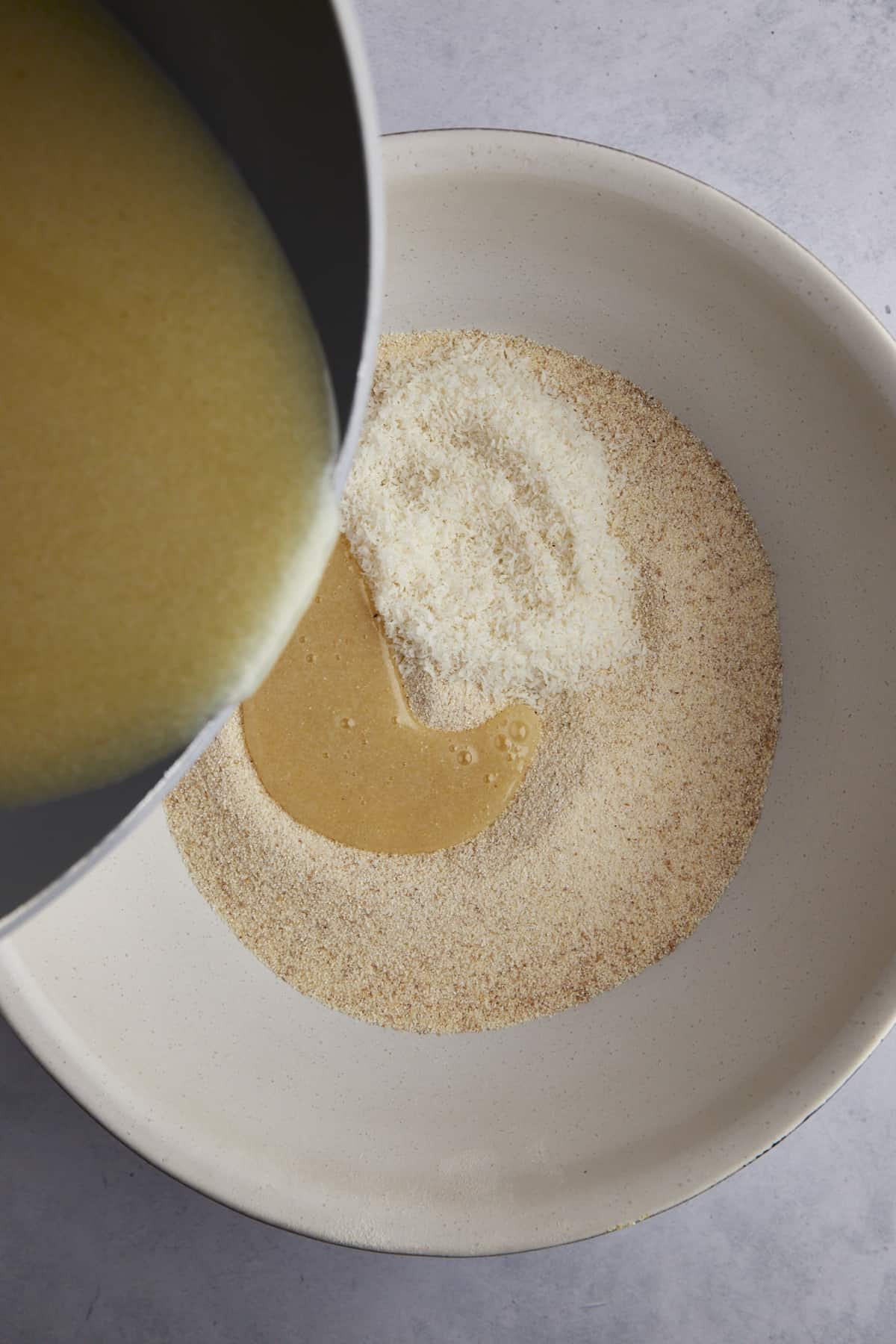 Melted butter being poured in a bowl of semolina flour and coconut flakes.