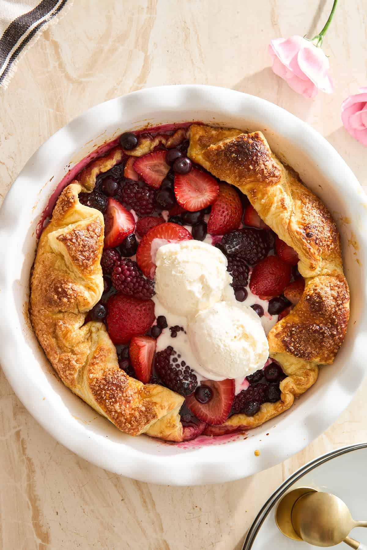A rustic mixed berry galette with golden crust, filled with strawberries, blueberries, and blackberries, topped with two scoops of vanilla ice cream, on a light marble surface with pink flowers nearby.