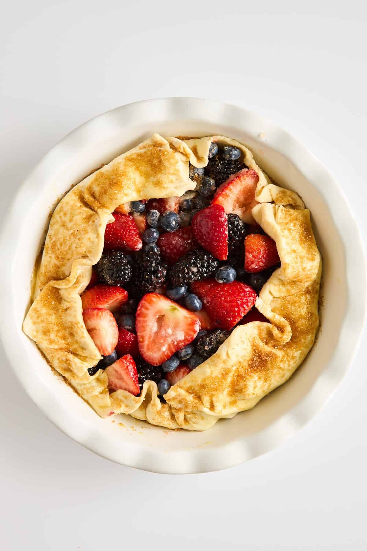 A rustic galette with a golden, folded crust filled with fresh strawberries, blueberries, and blackberries, sits in a white pie dish on a white background.