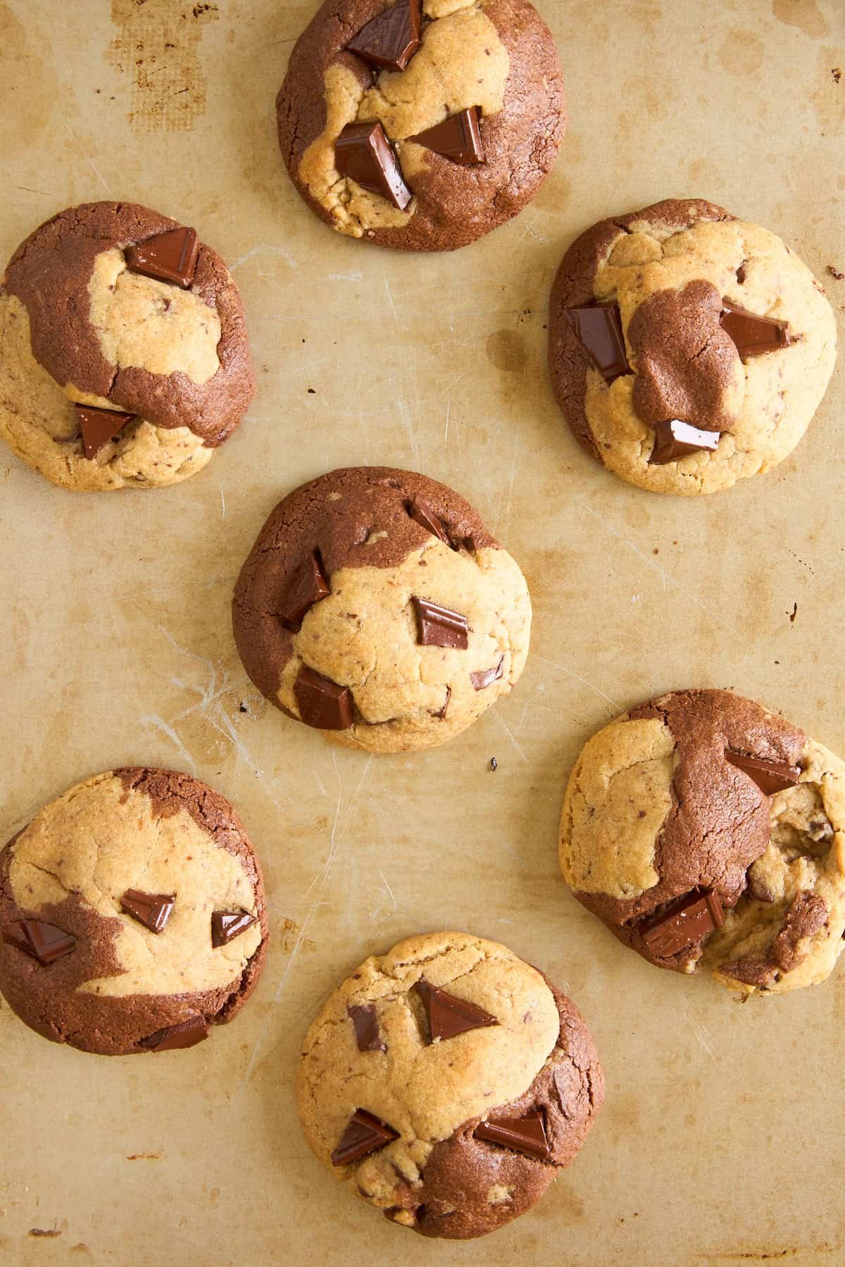 Seven large, marble-patterned cookies with chunks of chocolate are arranged on a tan baking sheet. The cookies have swirls of light and dark dough, and the surface is slightly cracked.