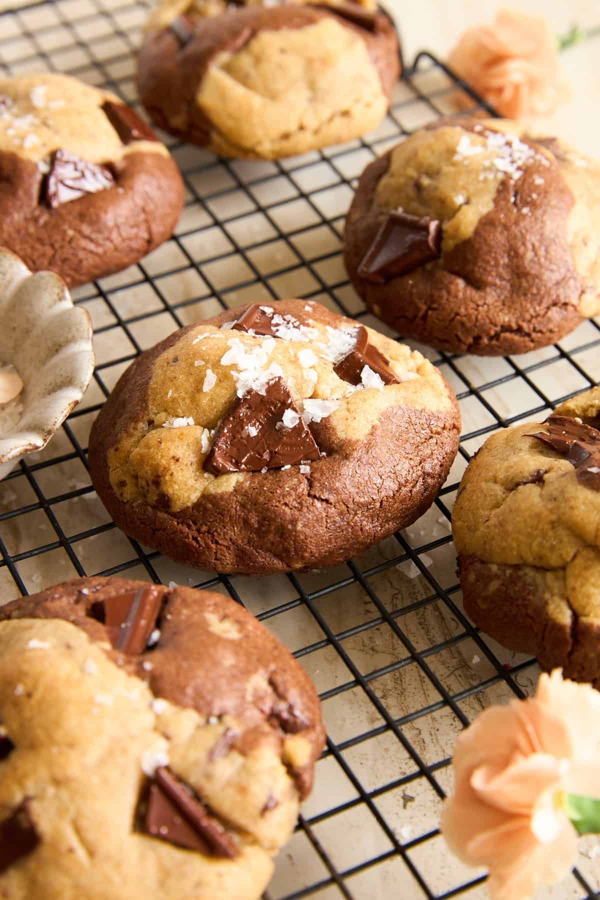 Chocolate chunk cookies with a marbled appearance rest on a black cooling rack. The cookies are sprinkled with flaky sea salt, and there are light orange flowers nearby for decoration.