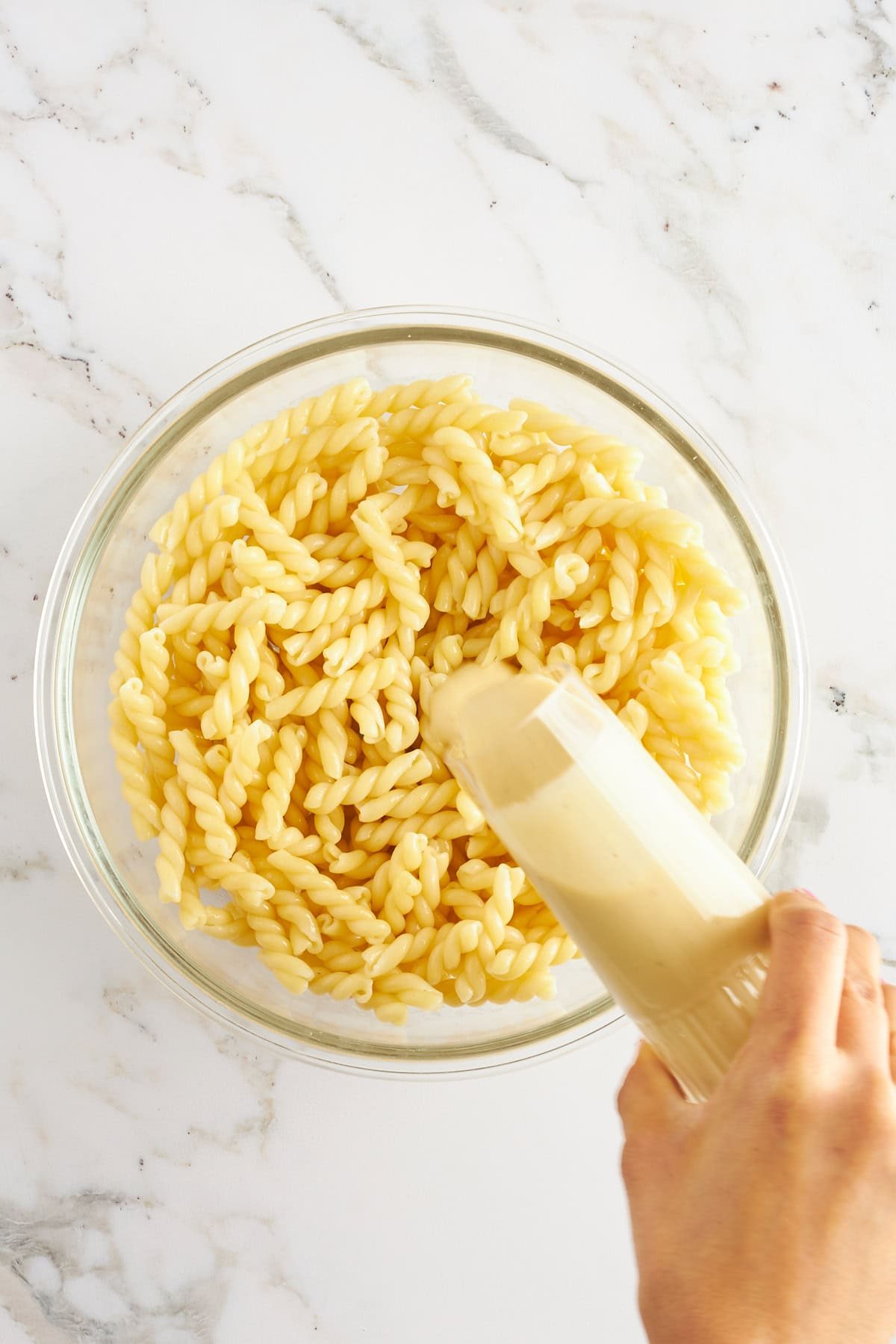 A bowl of medium-sized pasta noodles and a hand pouring Caesar dressing on top.