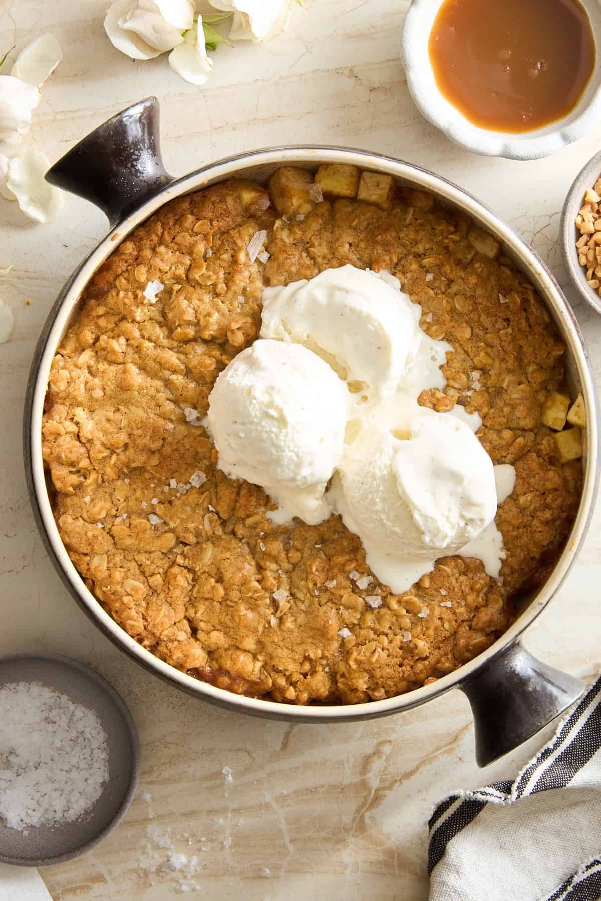 A round baking dish holds a golden-brown apple crisp topped with three scoops of vanilla ice cream. Nearby are bowls of caramel sauce, sea salt, and chopped nuts on a light marble surface.