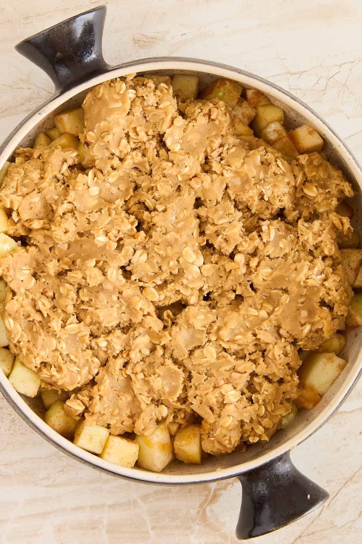 A round baking dish filled with apple chunks, topped with an uneven layer of oat crumble, ready to be baked. The dish has two black handles, and sits on a light-colored countertop.