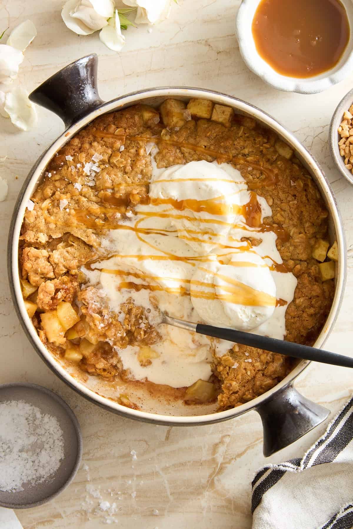 A round baking dish filled with apple crisp topped with melting vanilla ice cream and drizzled with caramel sauce. A spoon rests in the dish. Surrounding the dish are caramel sauce, nuts, a striped cloth, and flowers.
