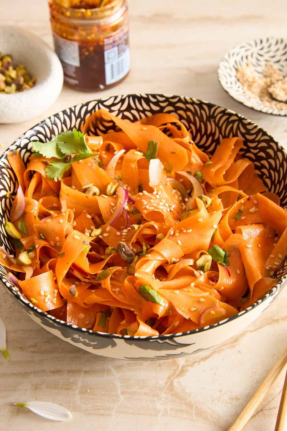 A patterned bowl filled with wide carrot ribbons, garnished with sesame seeds, chopped nuts, cilantro, and red onion slices, sits on a light surface with condiments and seeds nearby.