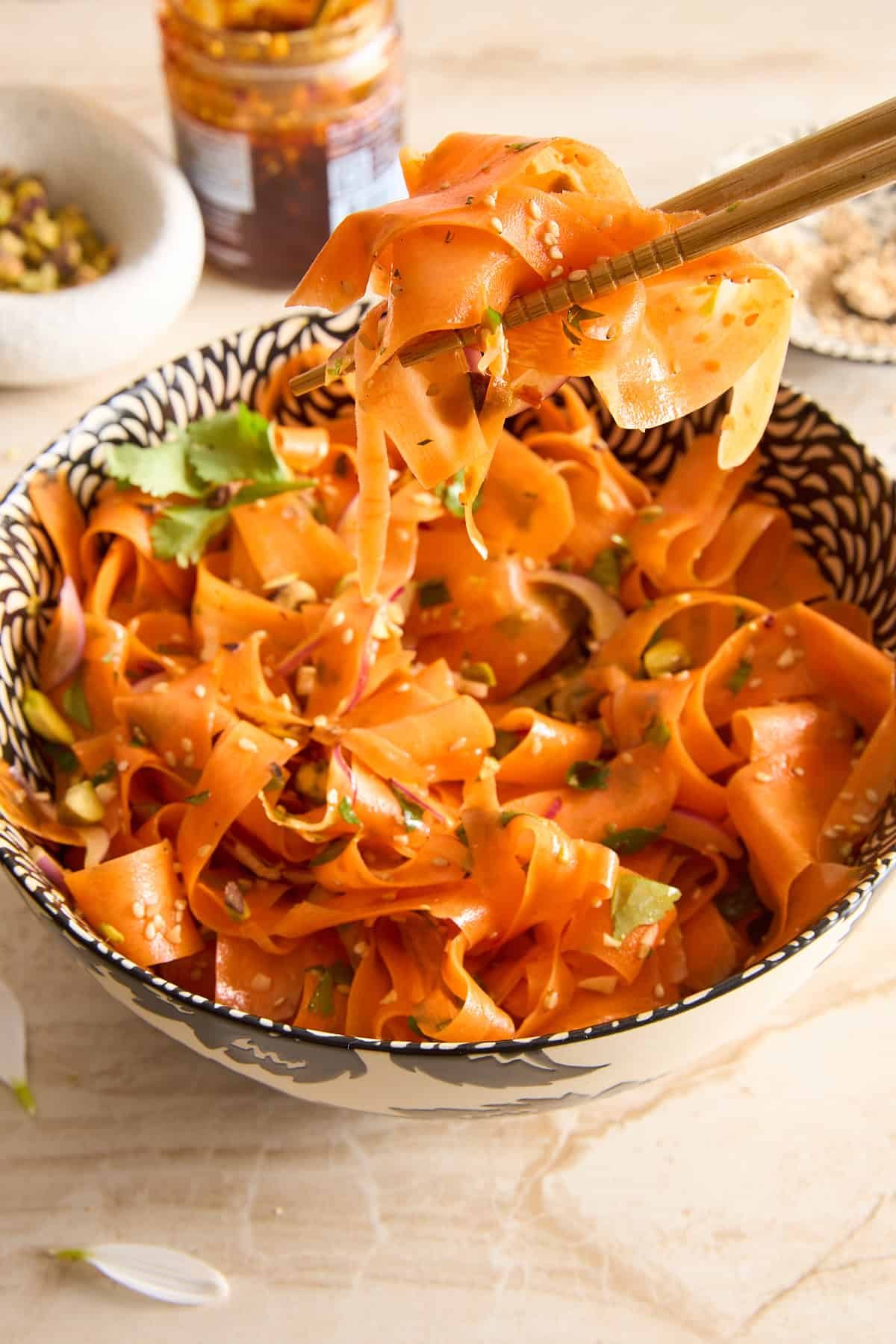 A patterned bowl filled with carrot ribbons garnished with herbs and seeds. Chopsticks hold up some carrot ribbons above the bowl. A jar of sauce and a small dish are visible in the background.