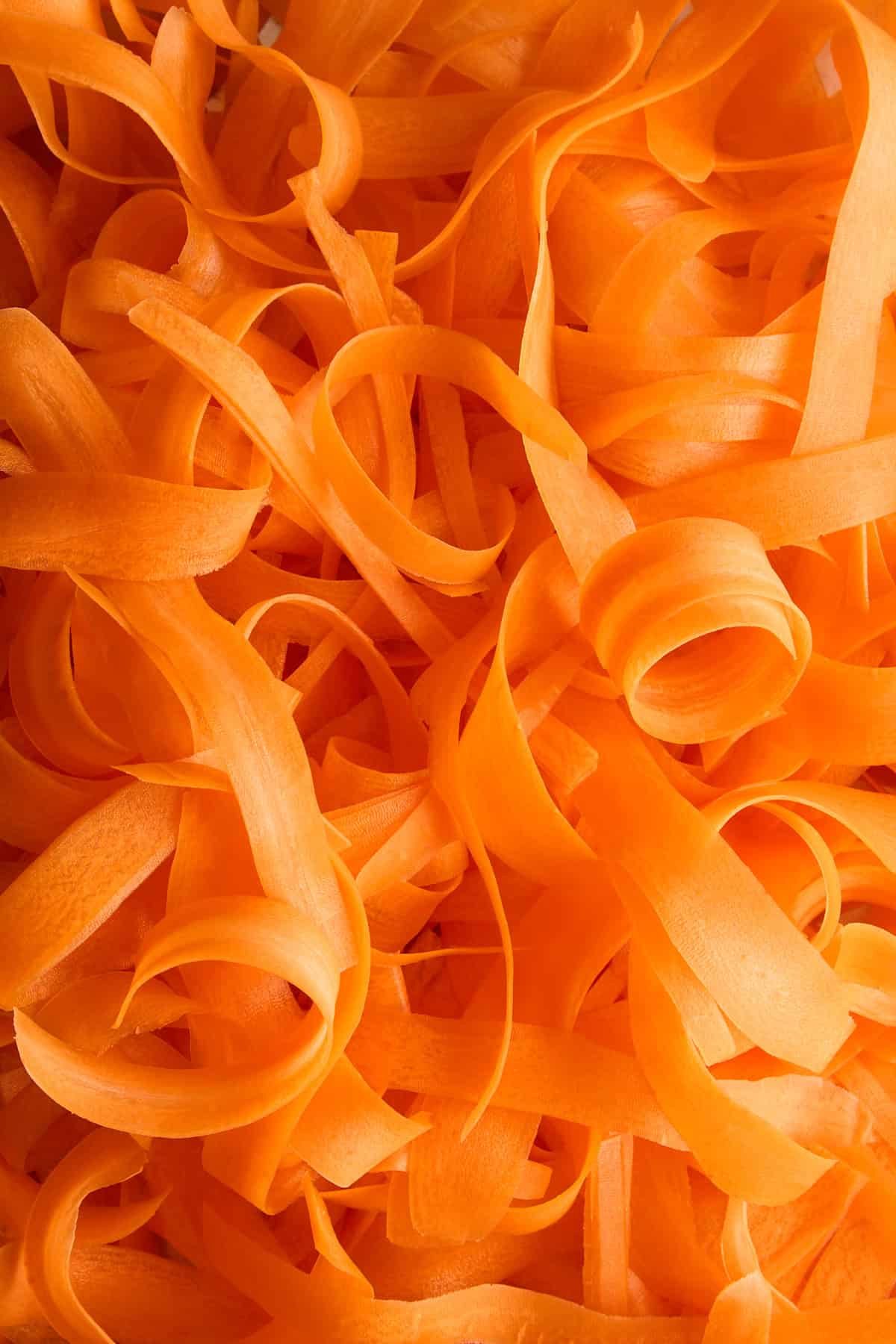 Thin, curly strips of bright orange carrot peel closely arranged in a random pattern, creating a textured and vibrant background.