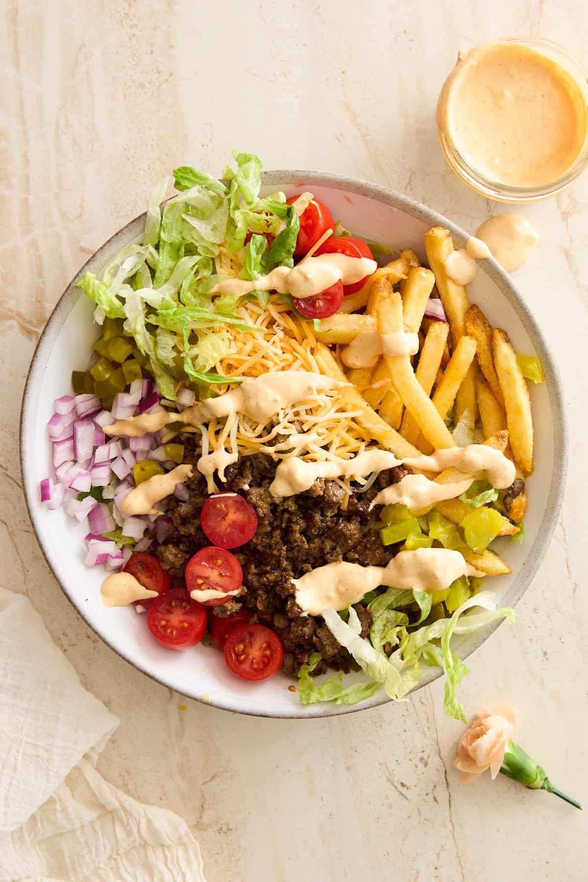 A bowl filled with shredded lettuce, sliced cherry tomatoes, shredded cheese, diced onions, pickles, ground beef, French fries, and drizzled with a creamy sauce. A small jar of sauce sits beside the bowl on a light surface.