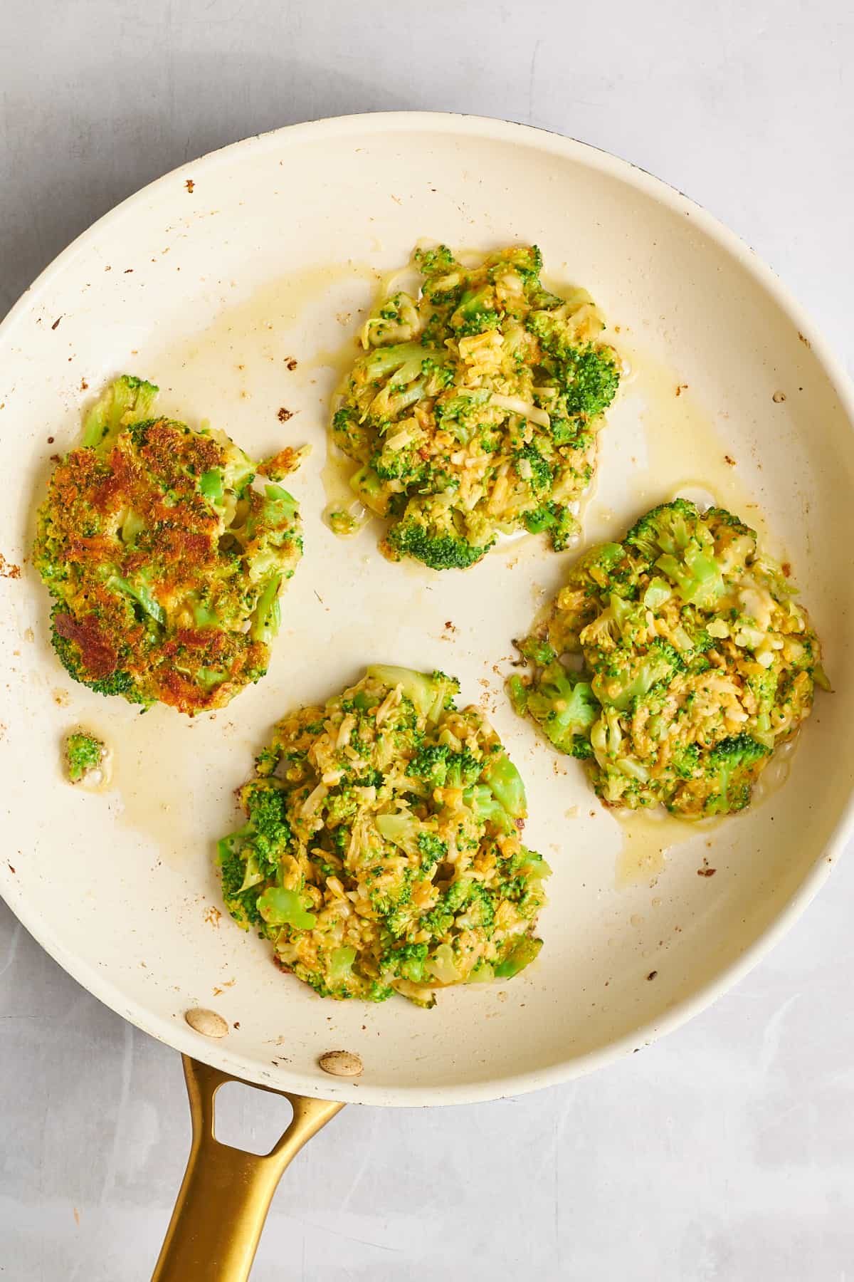 Four cheesy broccoli fritters cooking in a skillet.