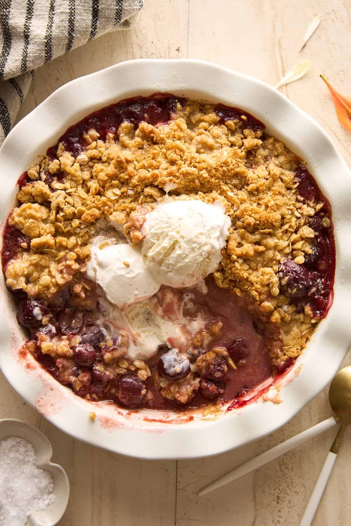 A baked cherry crisp in a white pie dish topped with two scoops of vanilla ice cream, with a portion served out, revealing the warm fruit filling and oat crumble topping. A spoon and fork are beside the dish.