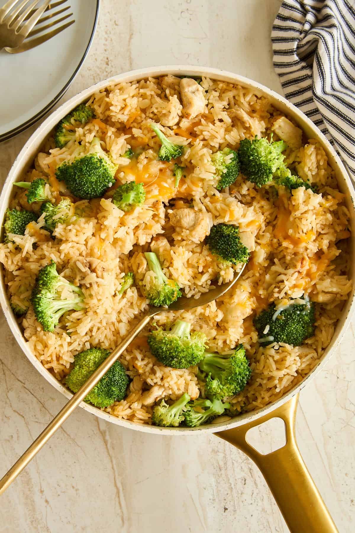 A skillet filled with rice, broccoli florets, and pieces of chicken, with a gold serving spoon resting inside. A striped cloth and stacked plates with forks are nearby on a light surface.