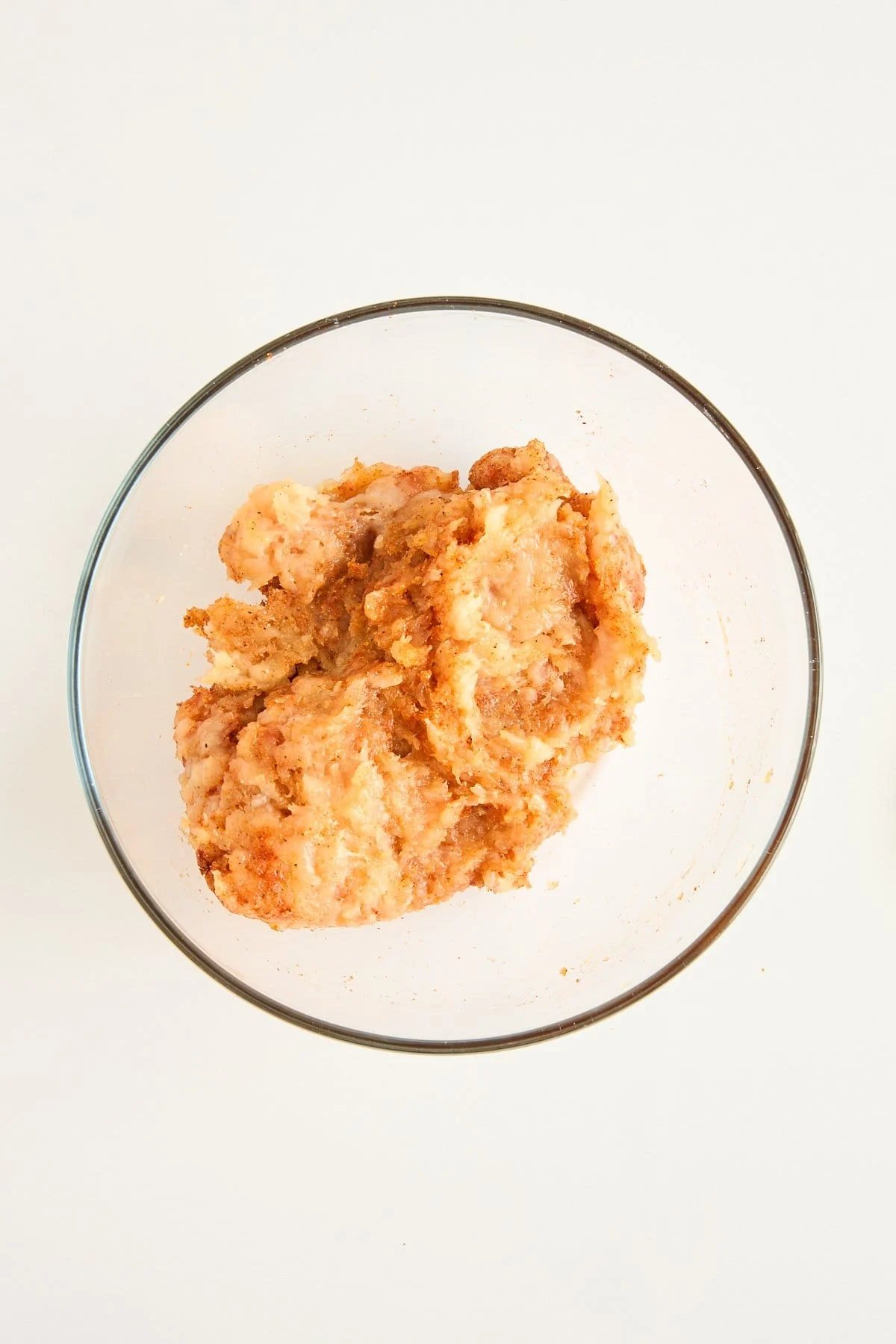 A clear glass bowl containing a mixture of ground meat and seasonings on a white surface.
