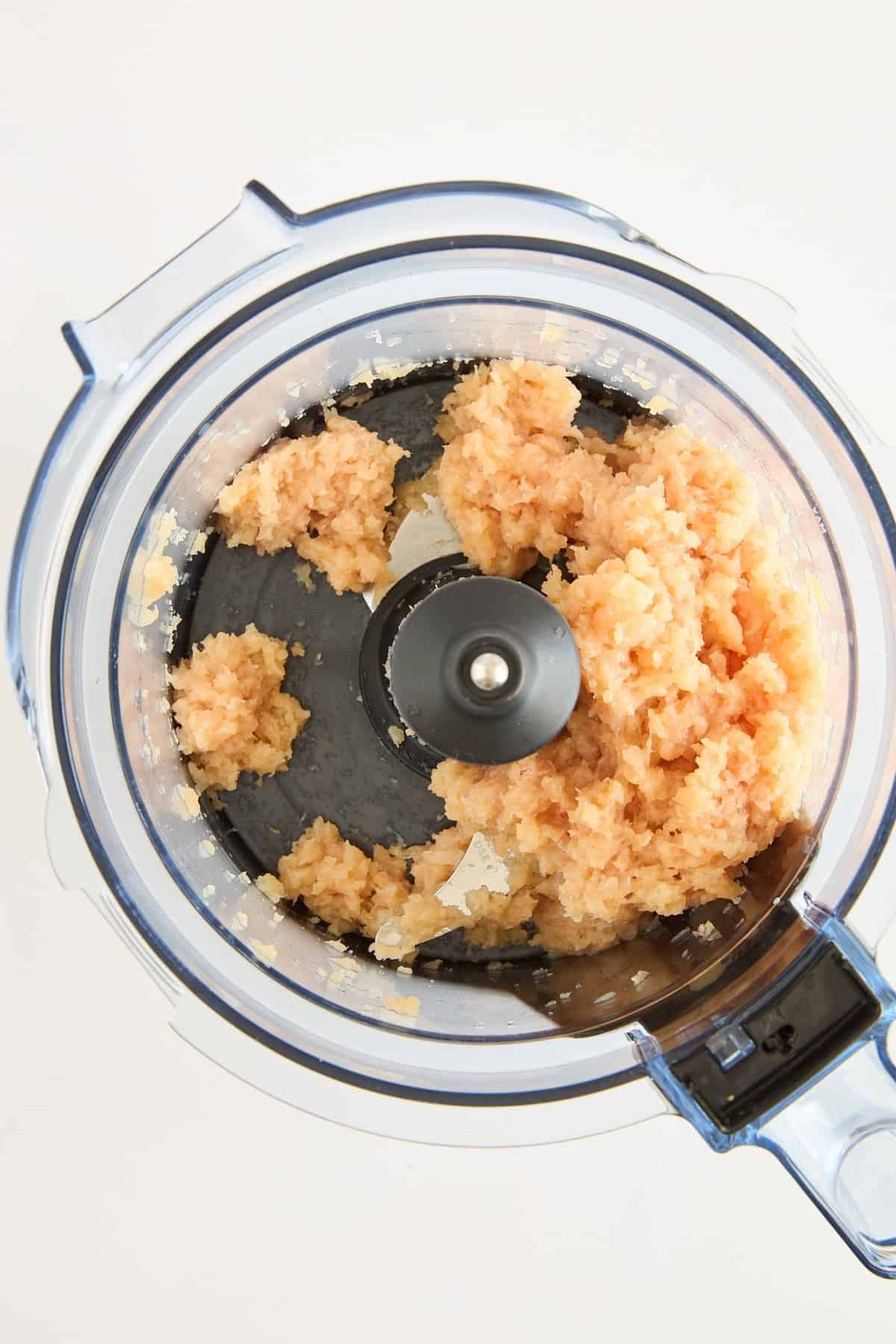 A top-down view of a food processor containing ground chicken and onions, partially blended, on a white background.