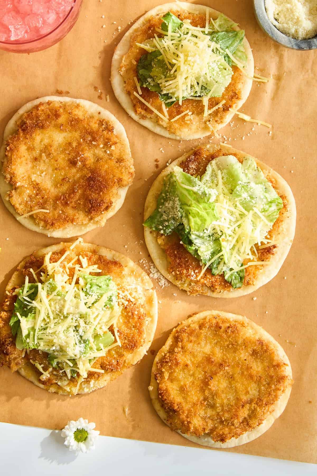 Five small round flatbreads are topped with breaded chicken, lettuce, shredded cheese, and grated parmesan, arranged on parchment paper with a small bowl of parmesan in the corner.