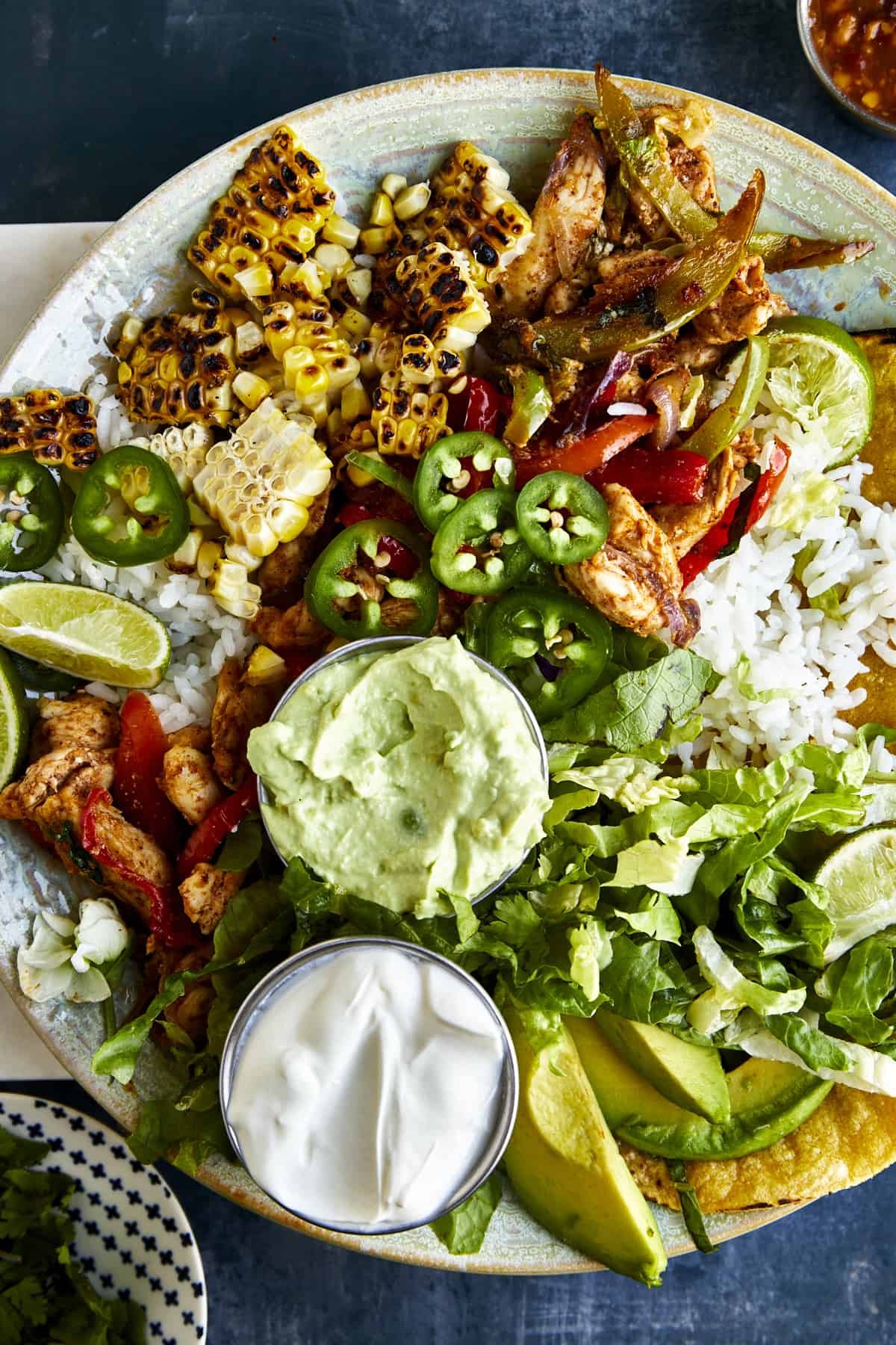 Overhead image of a chicken fajita bowl with rice, grilled corn, jalapeños, mashed avocado, sour cream, and corn tortillas.