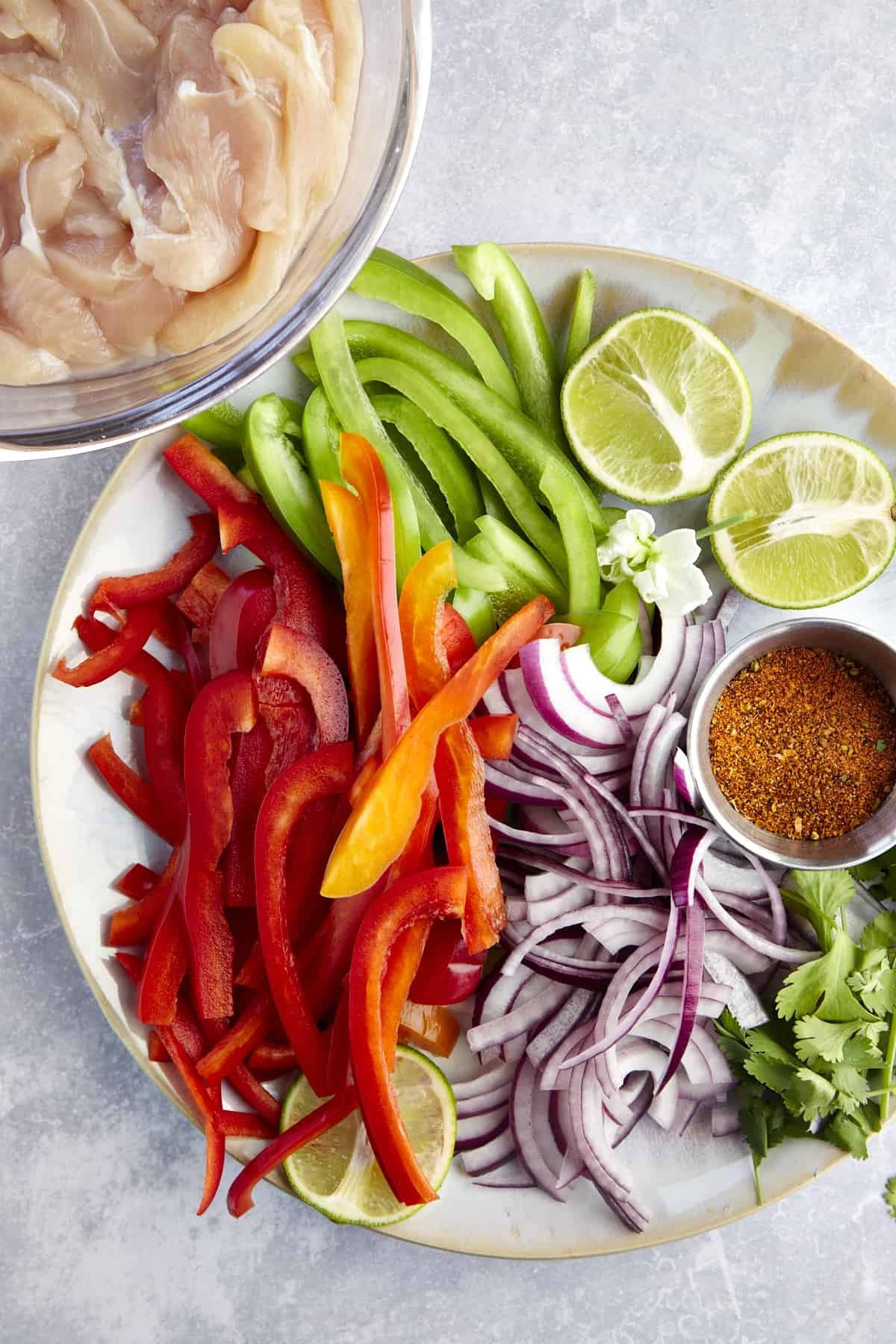 Sliced chicken, red and green bell peppers, red onion, a halved lime, cilantro, and fajita seasonings.