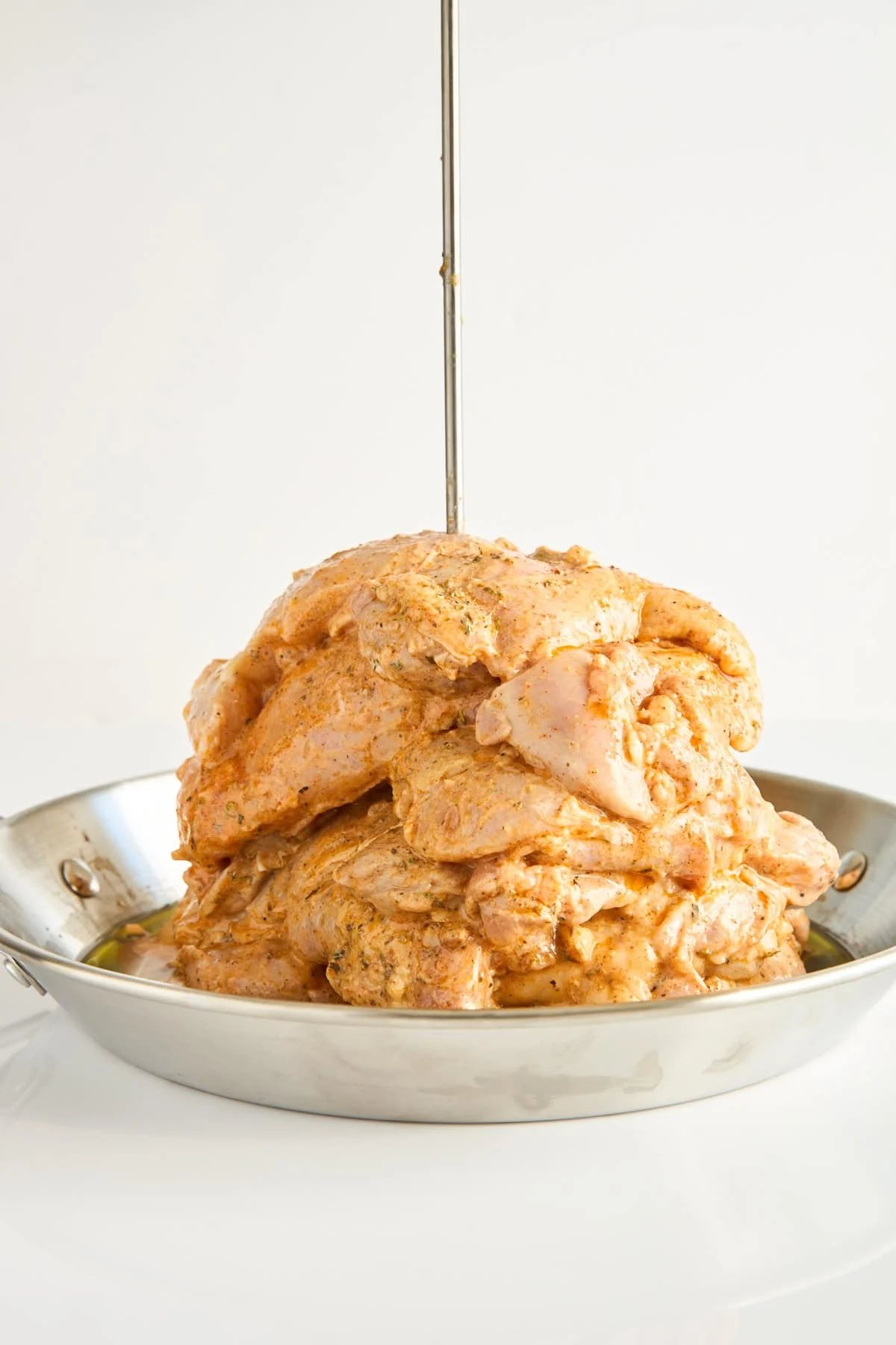 A stack of marinated raw chicken pieces is piled on a vertical skewer in a shallow metal pan, ready to be cooked, against a plain white background.