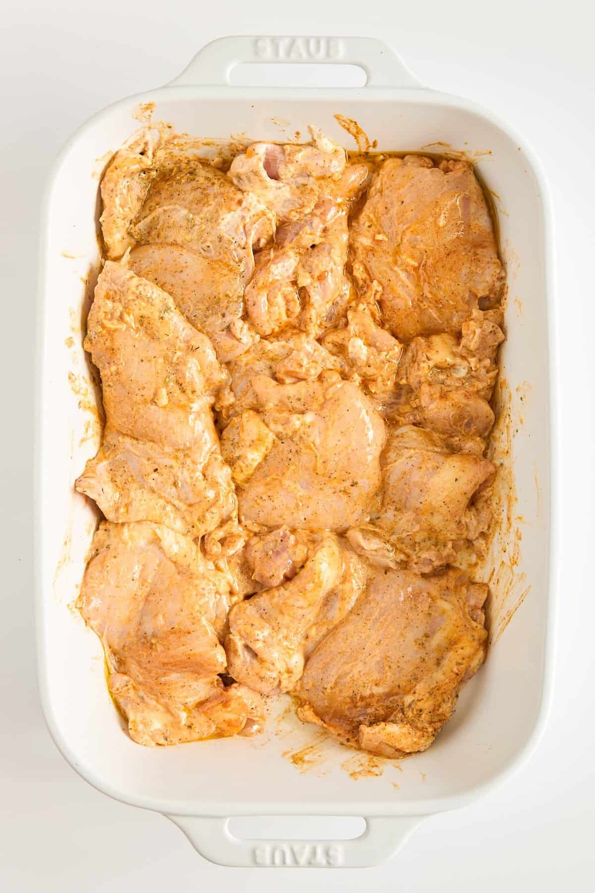Raw chicken pieces coated in a reddish-orange marinade are arranged in a single layer in a white rectangular baking dish. The chicken is uncooked and evenly distributed in the dish.