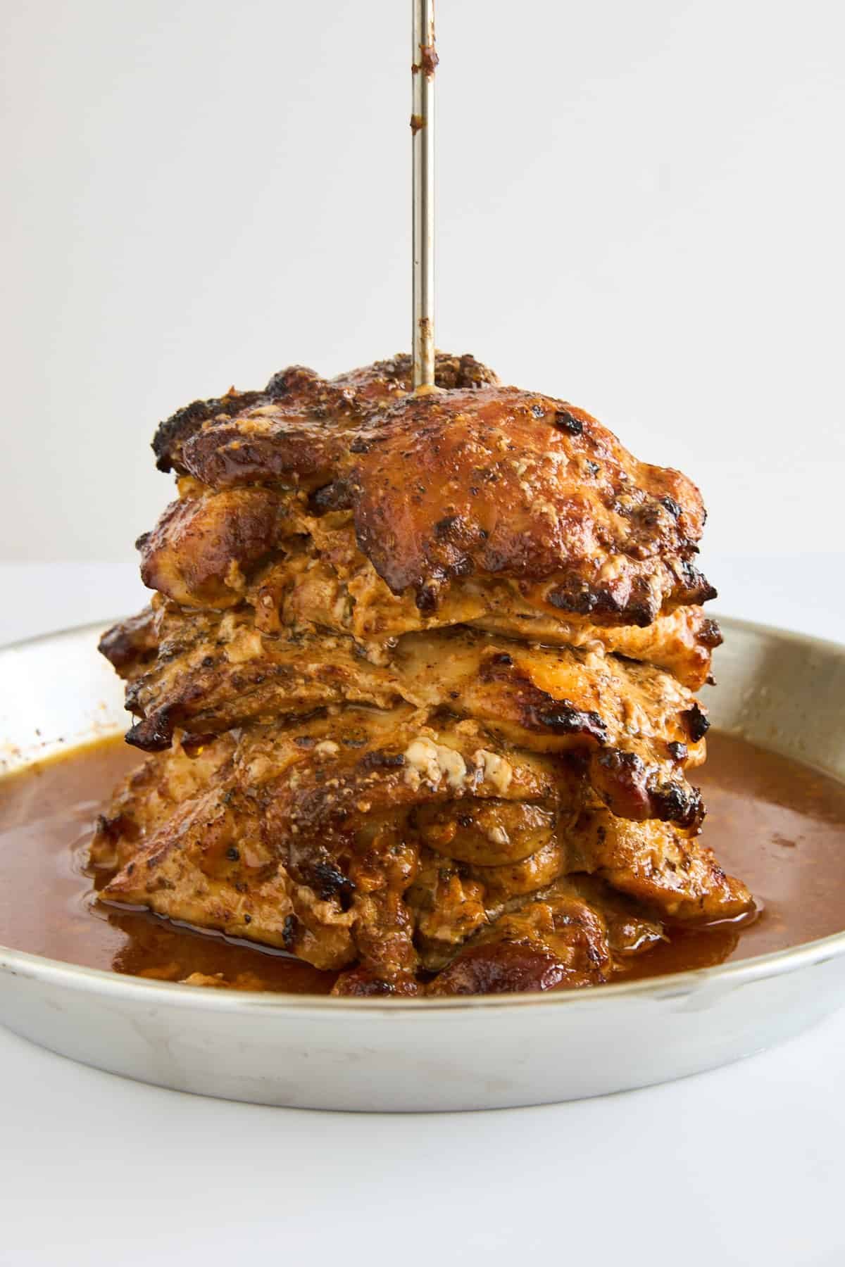 A stack of seasoned, roasted meat slices skewered vertically on a metal rod, sitting on a round metal tray with juices collecting at the bottom.