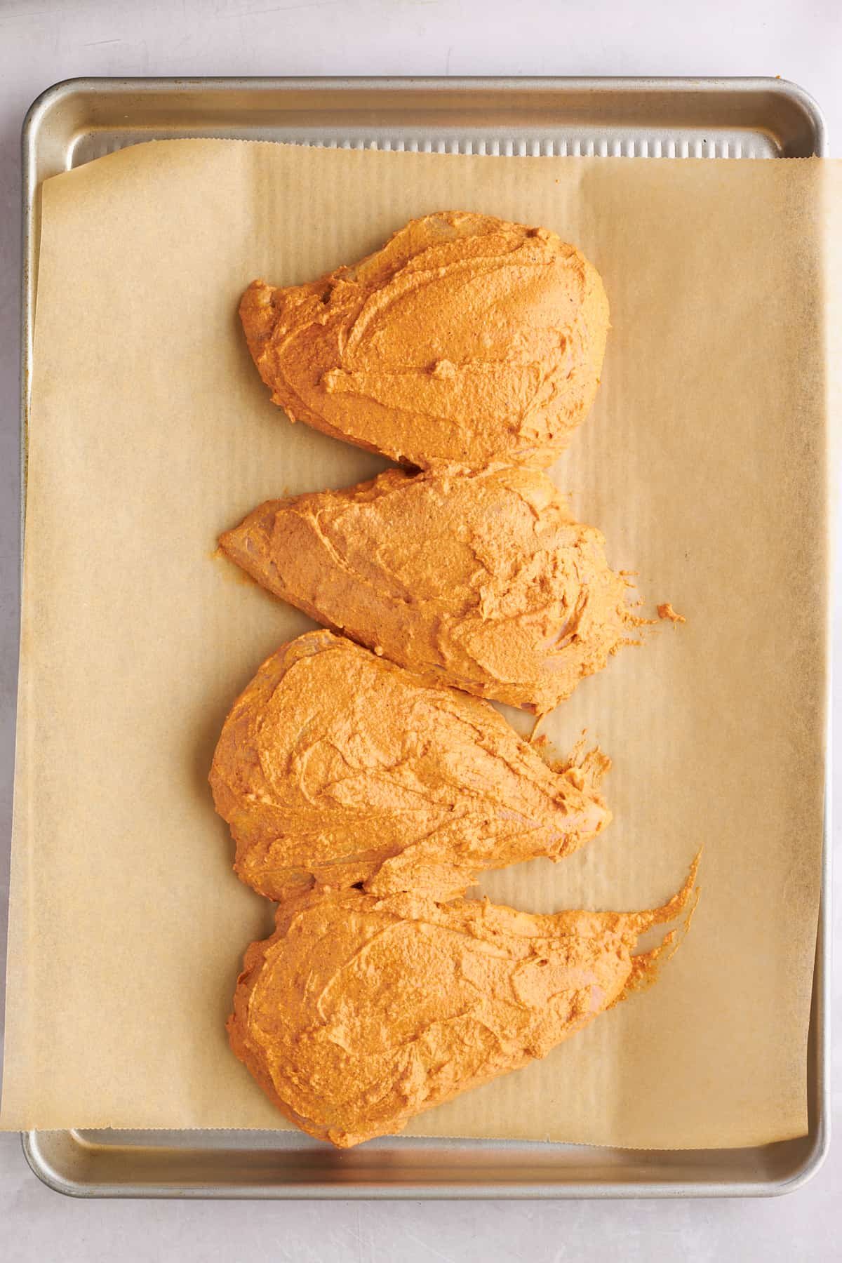 Four marinaded chicken breasts on a parchment paper-lined baking sheet.