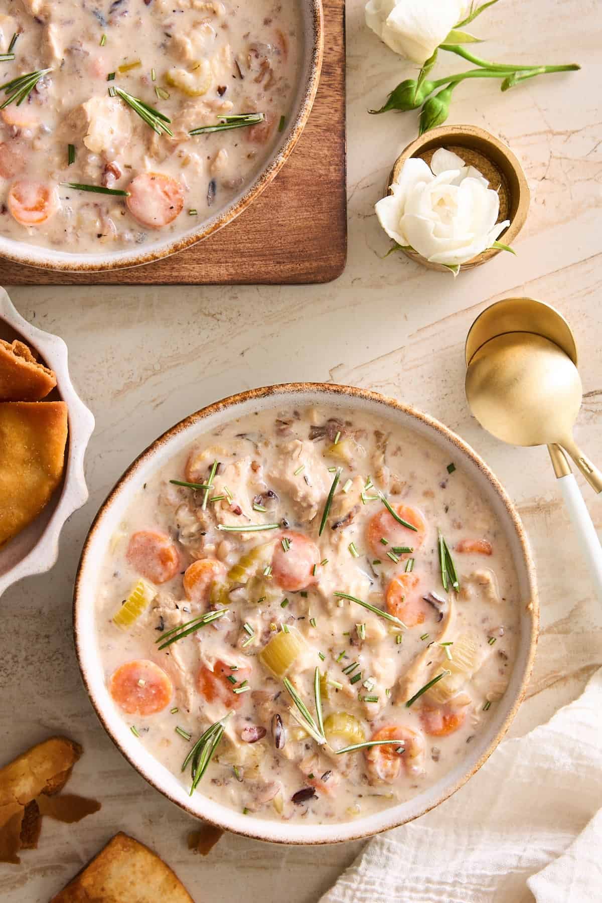 A bowl of silky soup with carrots, celery, wild rice, and herbs sits on a light table next to a gold spoon, white flowers, toasted bread, and another bowl on a wooden board.