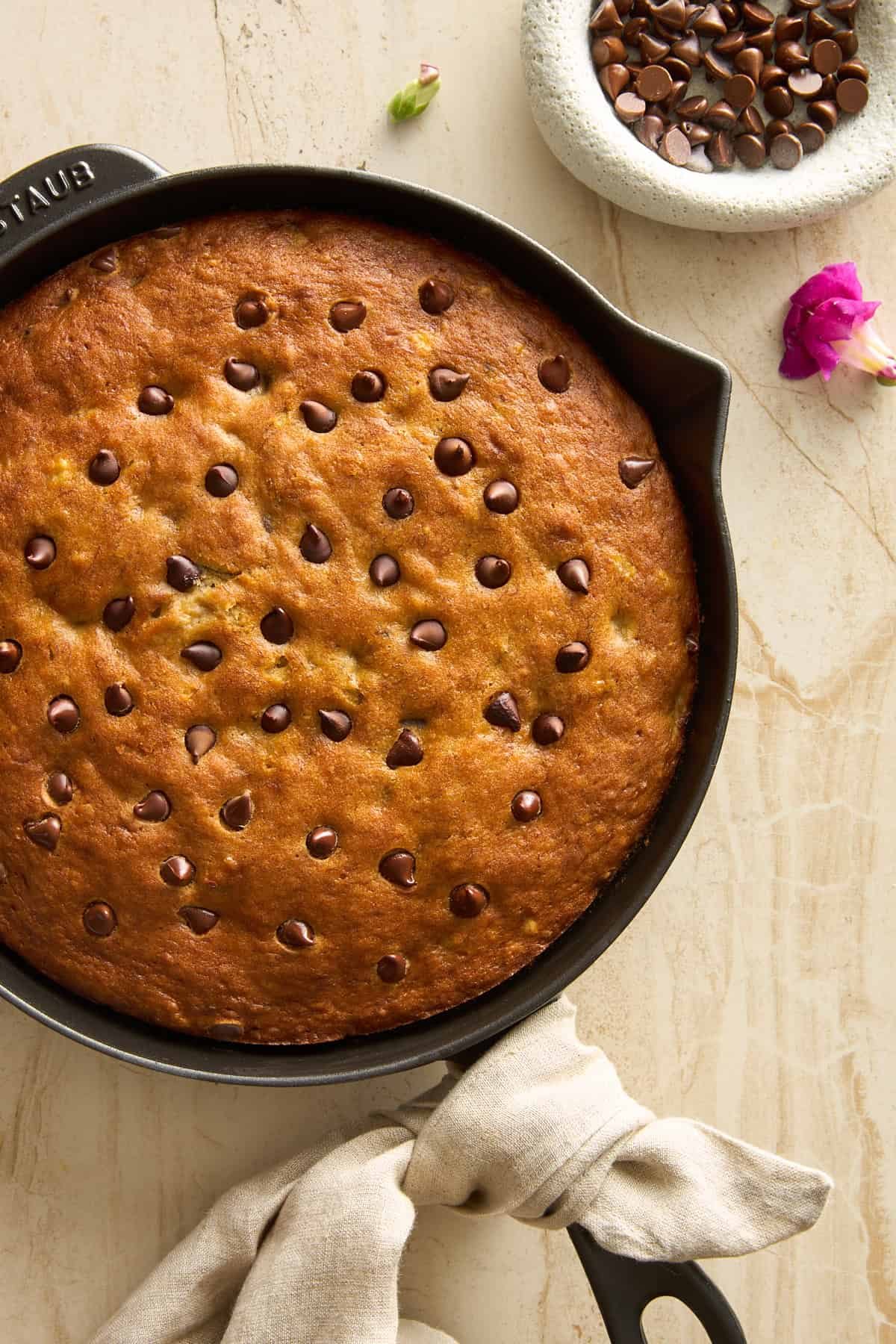 A skillet of freshly baked banana bread topped with chocolate chips sits on a light-colored surface. A beige cloth is wrapped around the skillet handle, and a bowl of chocolate chips and flower petals are nearby.