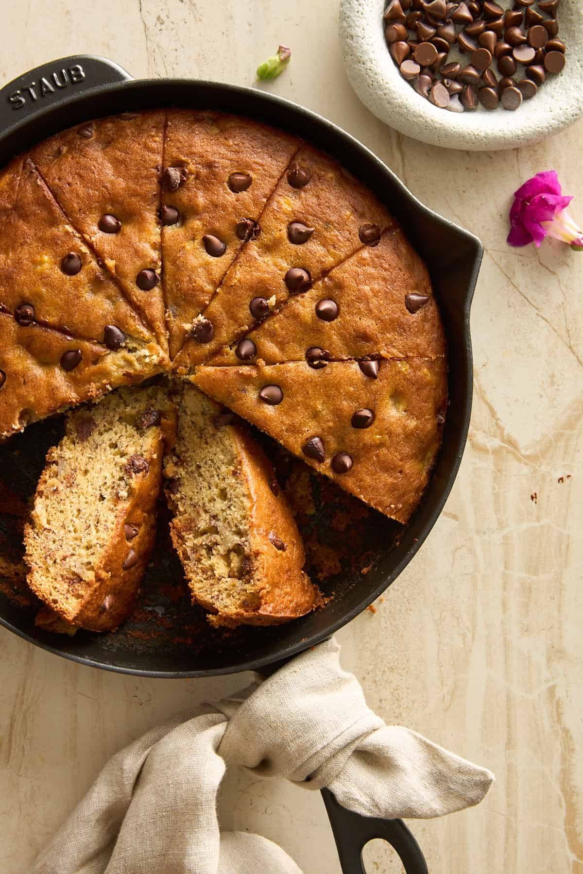 A round chocolate chip banana bread in a cast iron skillet, sliced with two pieces slightly removed. A bowl of chocolate chips and a pink flower are nearby on a light surface. A beige cloth wraps the skillet handle.